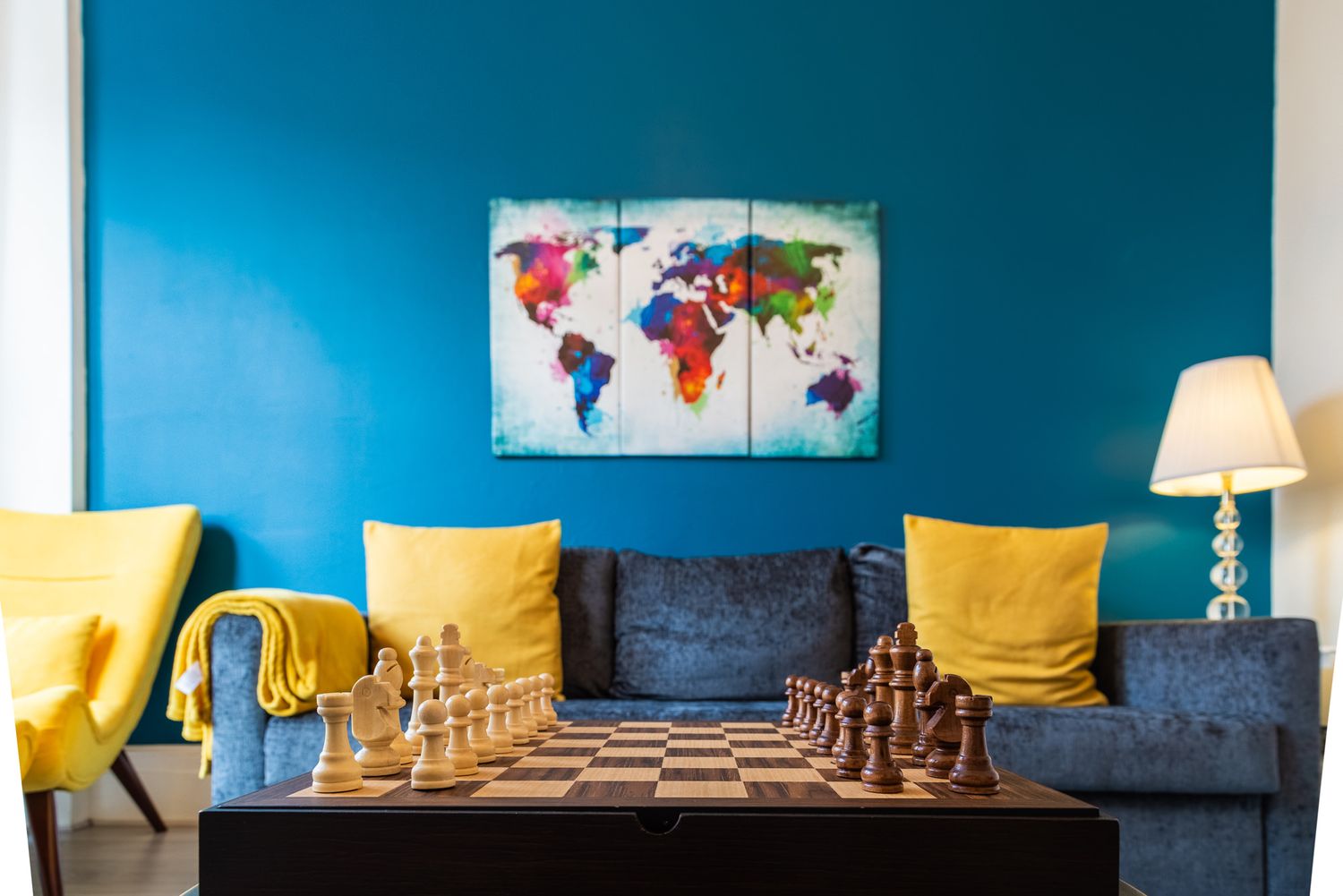 A modern living room with blue walls, yellow throw pillows, chess set on gray couch, and colorful world map artwork.
