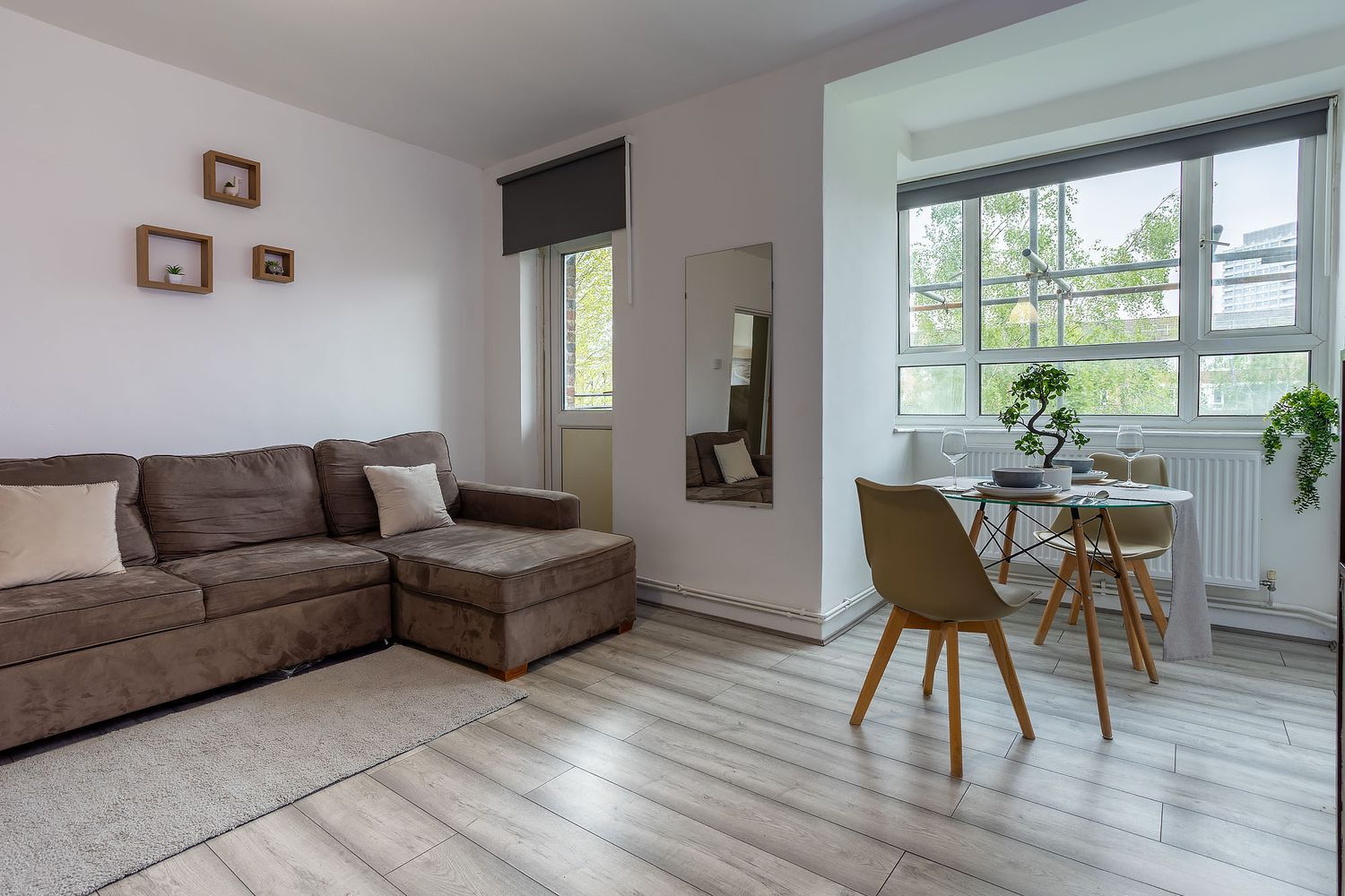Modern apartment interior with brown sectional sofa, light wood flooring, and dining nook near bright windows with plants.
