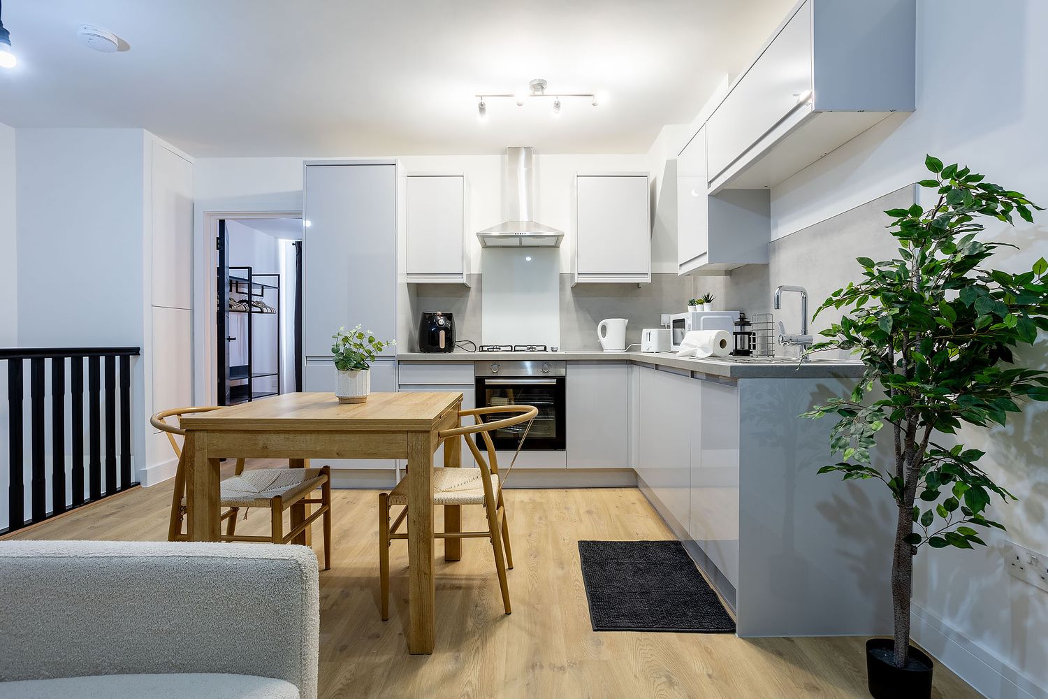 Modern white kitchen with light wood dining table, hardwood floors, and decorative indoor plant in open concept space.