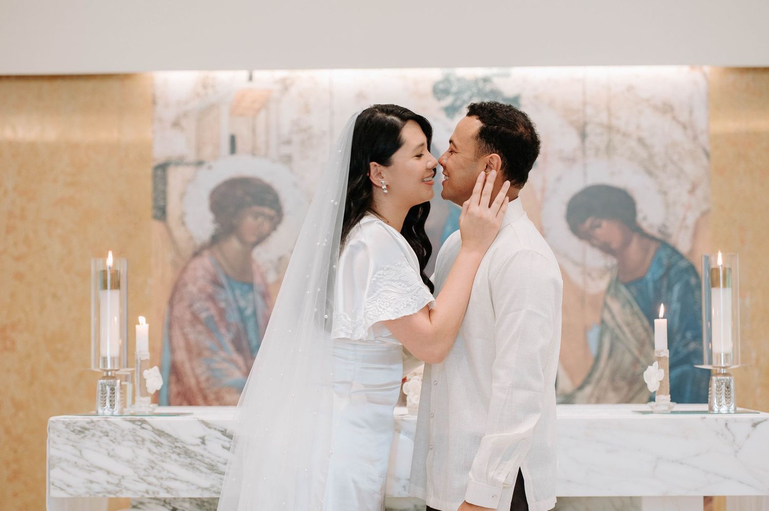 A couple in white outfits share a romantic moment at a church altar with religious artwork and candles in the background.