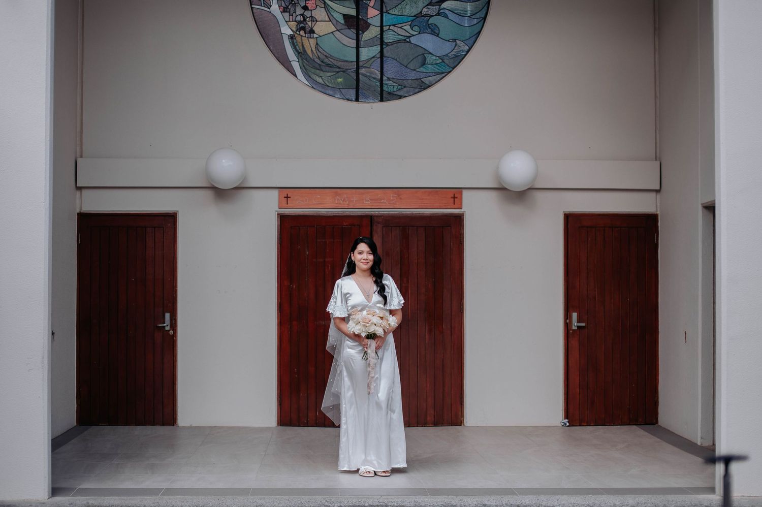 Someone in a white dress stands holding flowers in front of wooden doors with a circular window above.
