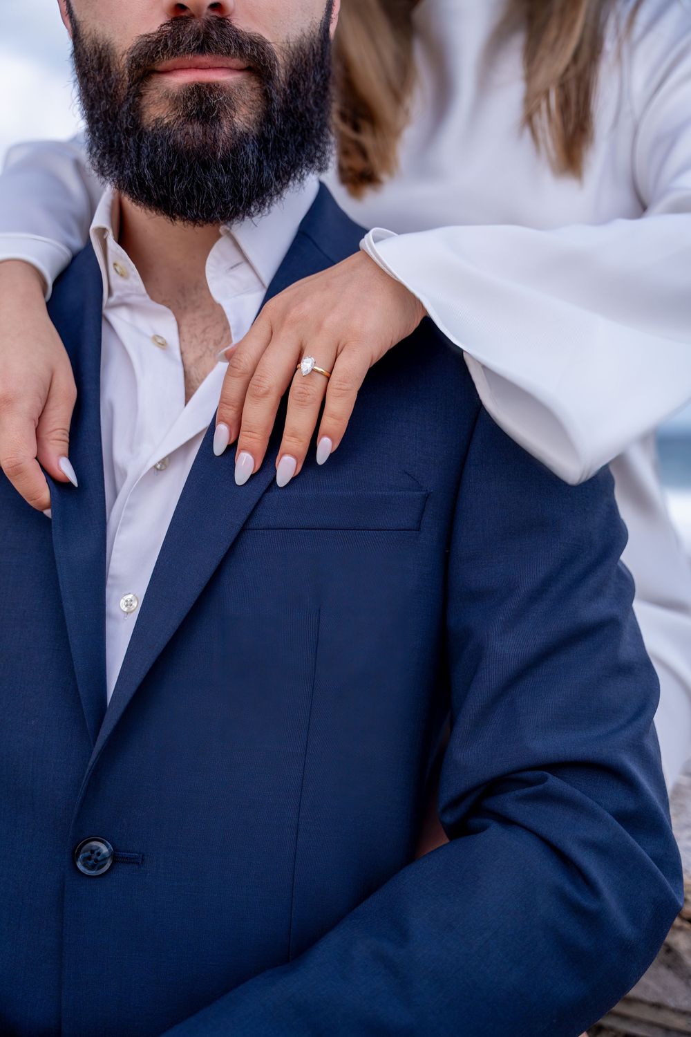 Close-up of an engagement ring on hand resting against a navy blue suit jacket.