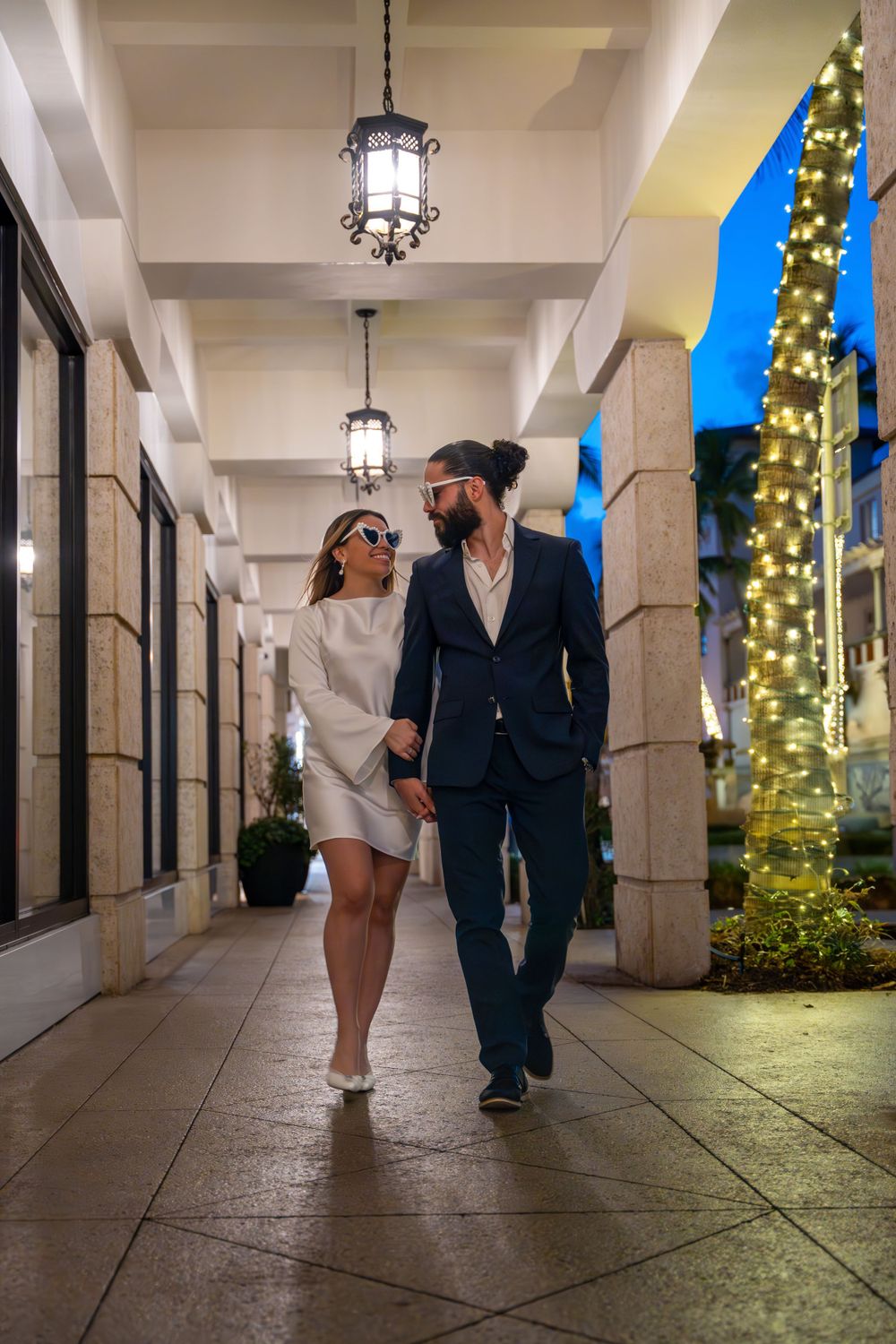 Stylishly dressed couple walking under illuminated archway with decorative lights at night.