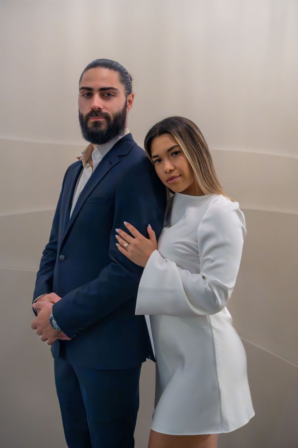 Couple in contrasting navy suit and white dress pose together against neutral wall.