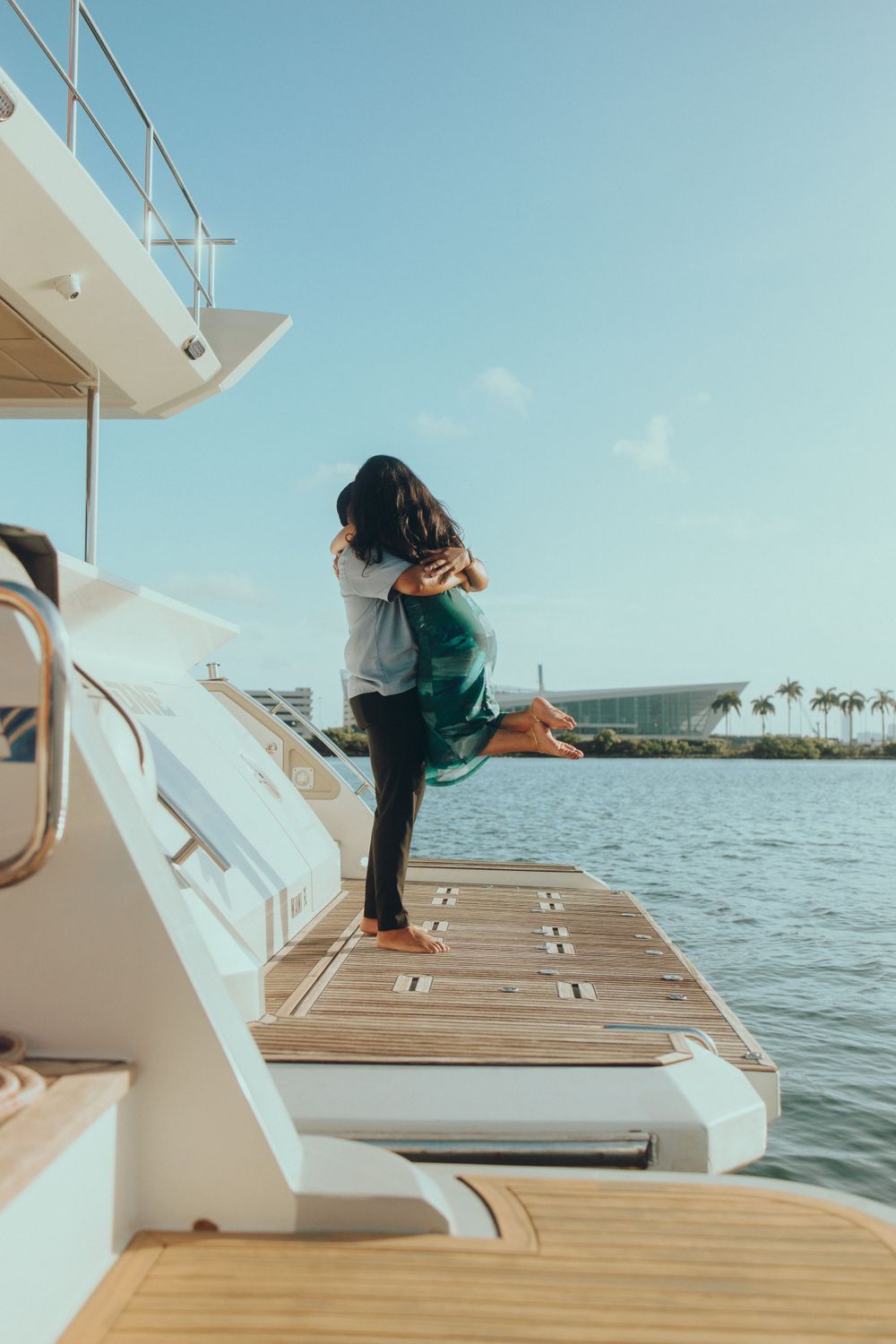 Romantic embrace after proposal on yacht deck with marina and palm trees in background.