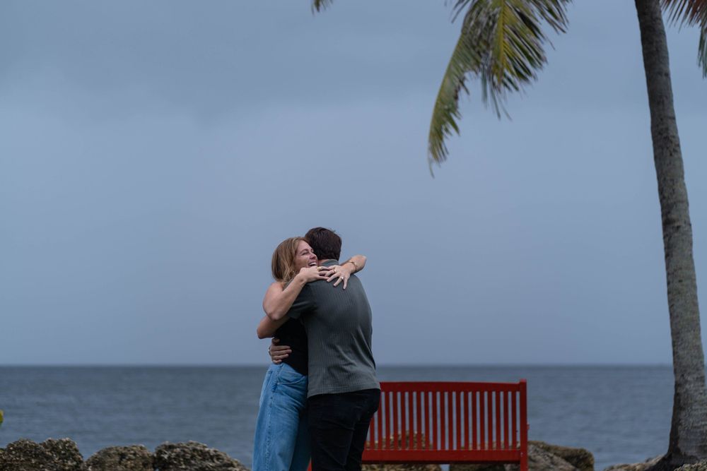 Couple hugging near a red bench with the ocean and palm trees in the background at The Kampong, Miami.