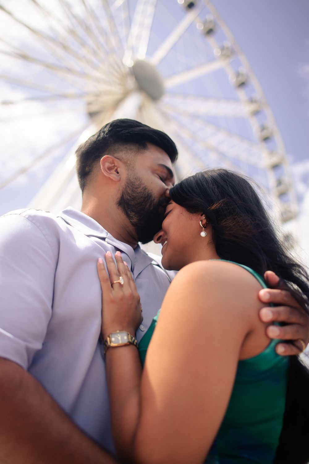 Engaged couple sharing intimate moment in front of Ferris wheel on sunny day.