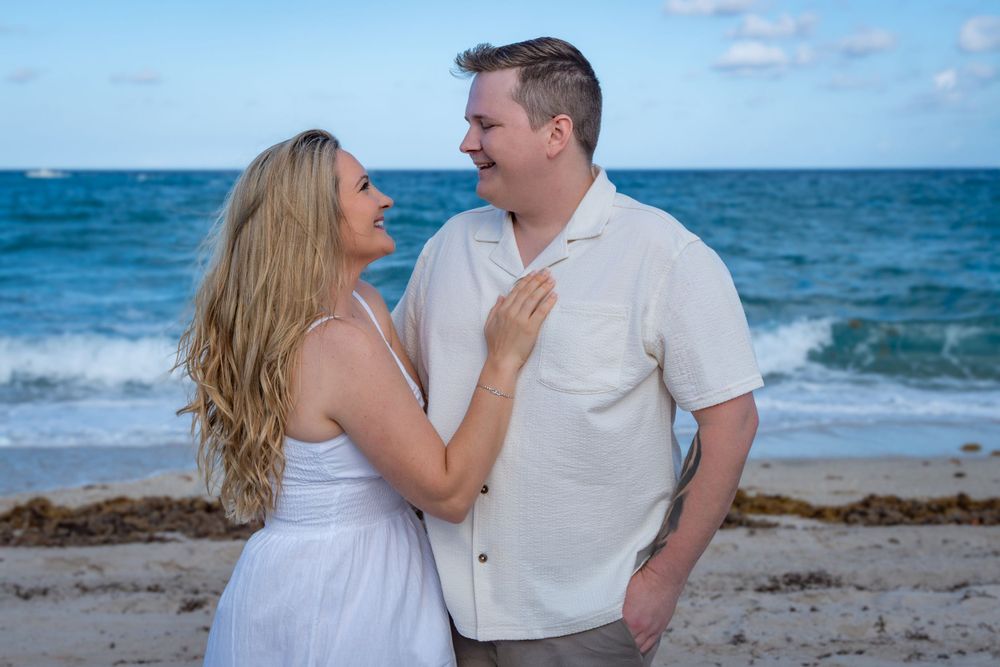 Couple in white outfits sharing romantic moment on beach with ocean backdrop.