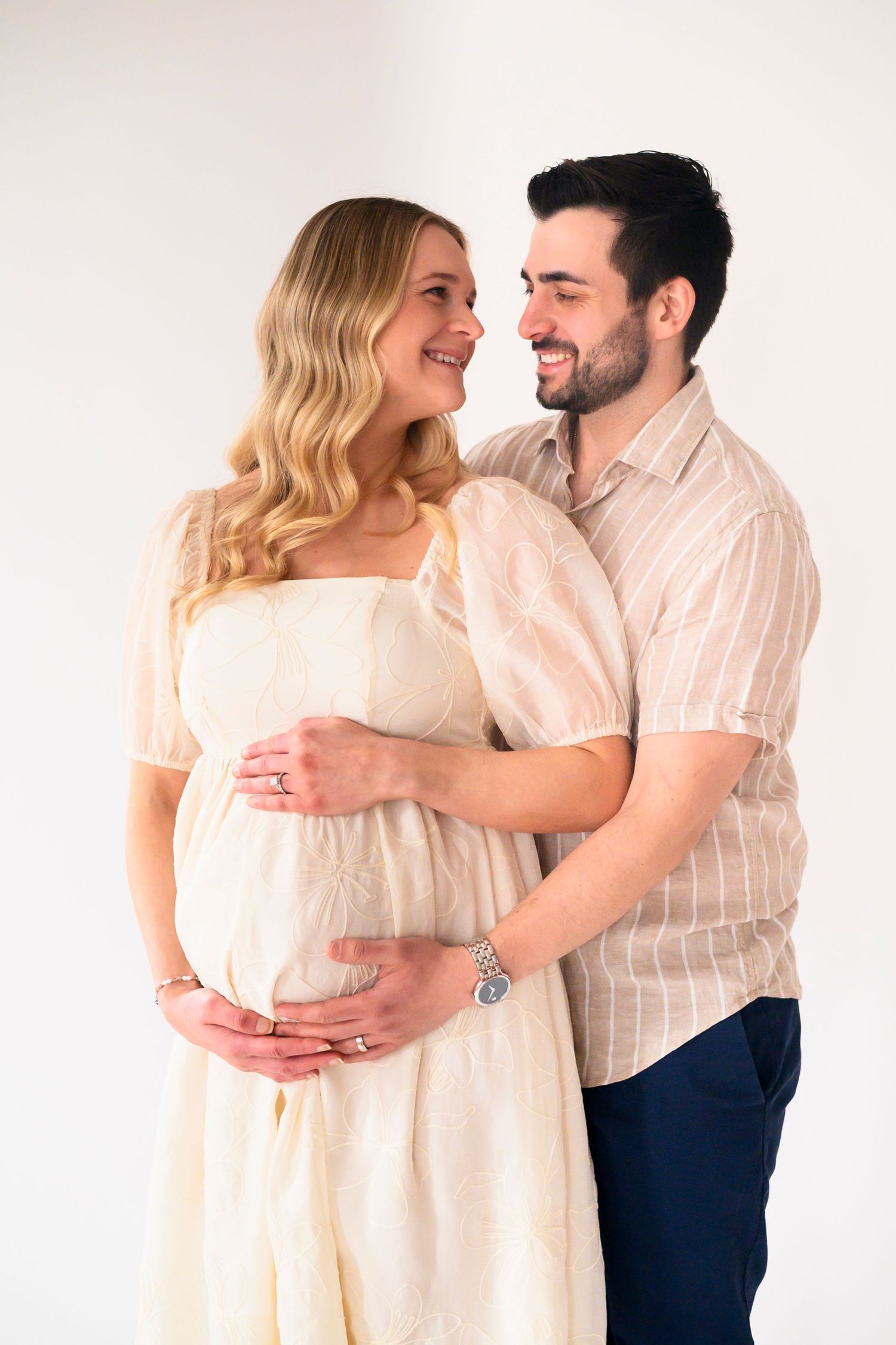 An expectant couple shares a tender moment against a white backdrop, wearing light colored clothing while embracing.