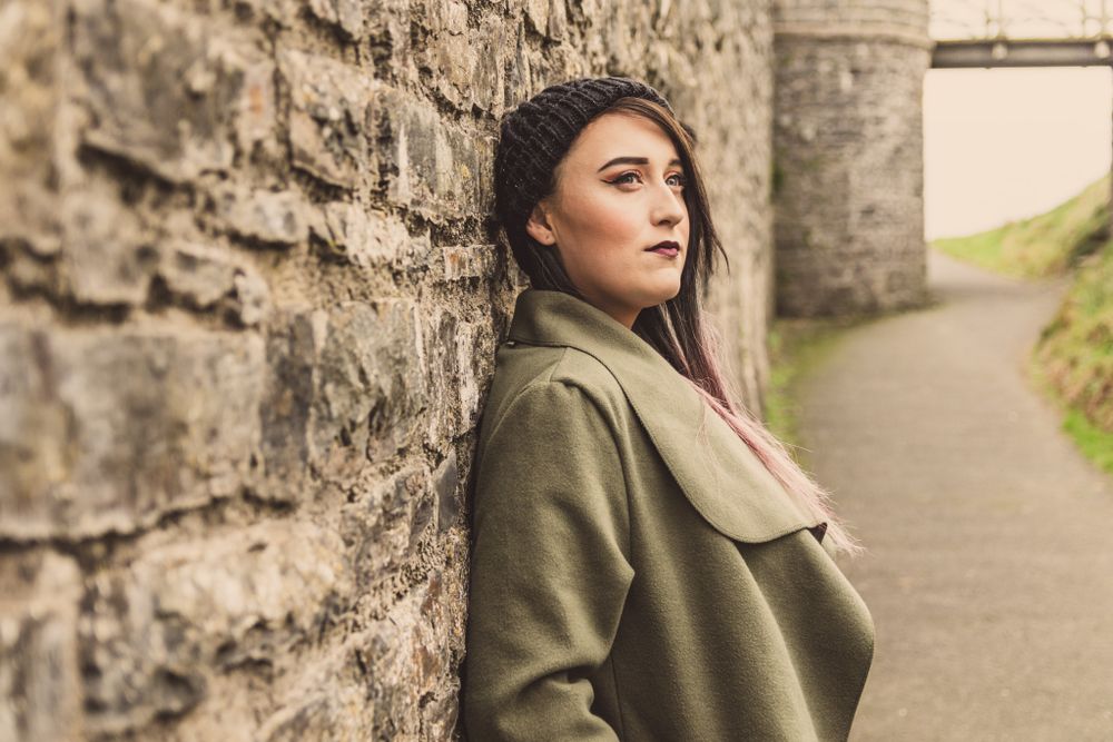 A portrait of a young lady with long dark hair, in a green coat leaning against an Aberystwyth castle wall. Wales Portrait Photograher