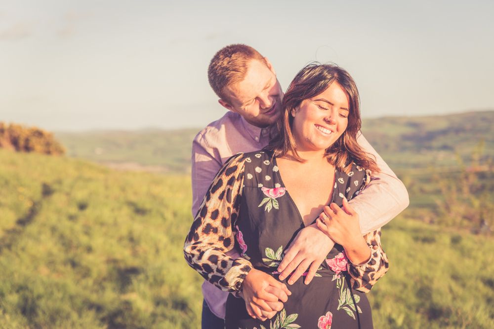 A portrait of a loving couple at golden hour in summer. He's hugging her from behind while they are standing. Conwy Portrait Photographer