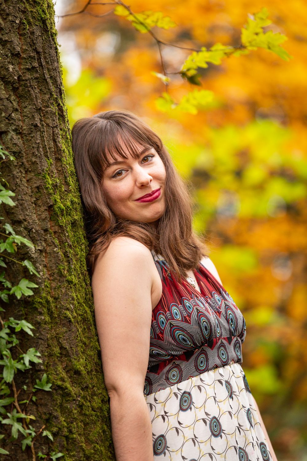 A portrait of a young woman in a sleeveless dress leaning against a tree with Autumn leaves behind. North Wales Portrait Photographer