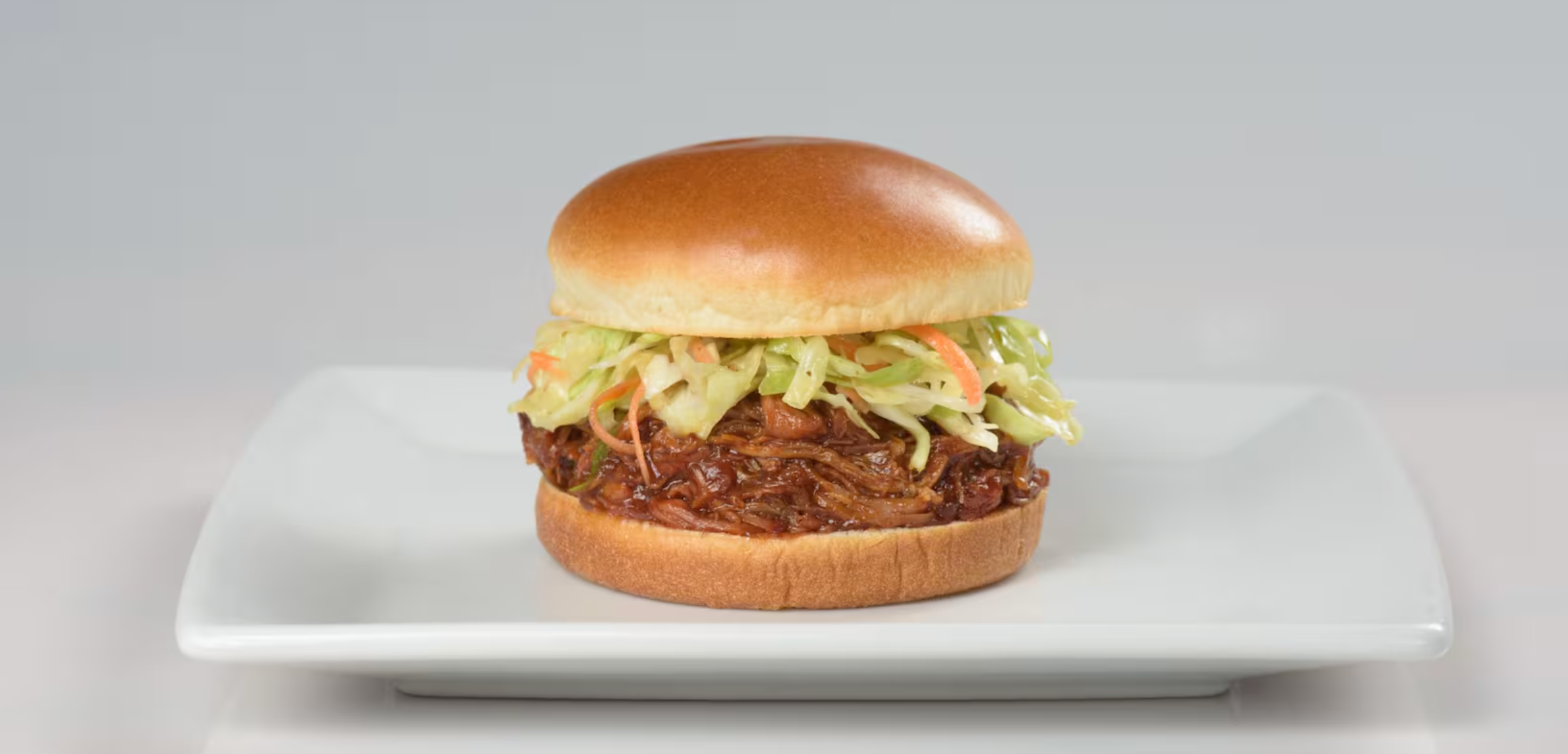 A pulled pork sandwich with coleslaw on a brioche bun sits centered on a white square plate.