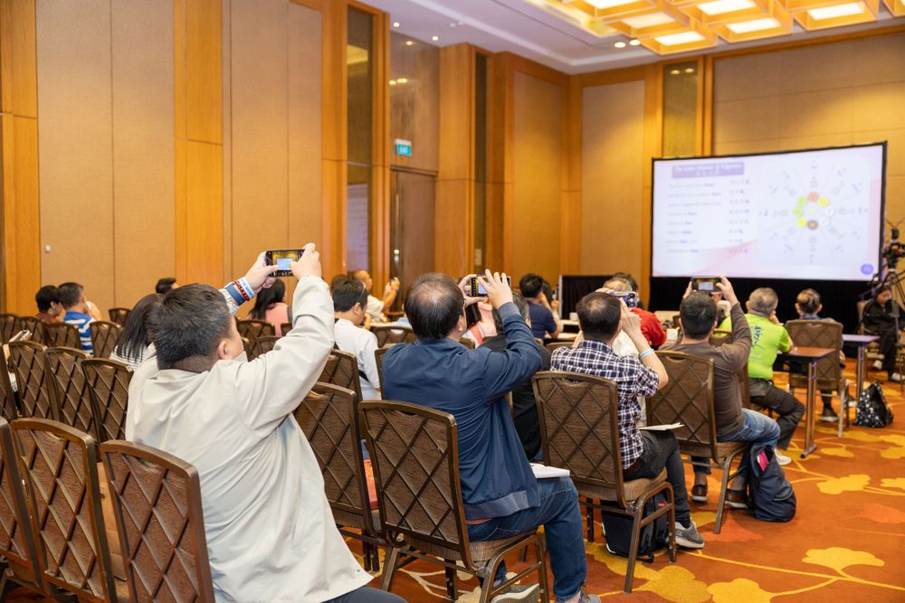 Conference attendees raising hands during interactive presentation session in hotel ballroom.