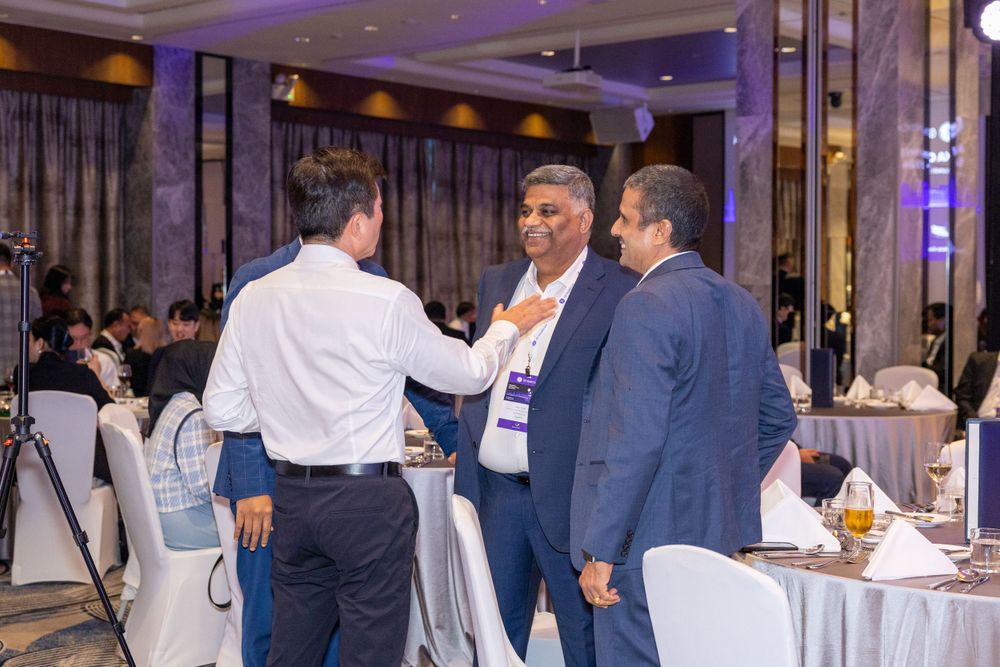 Business professionals engage in conversation at formal event in elegant hotel ballroom setting.