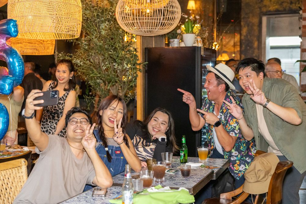Group of diners celebrate enthusiastically at a restaurant table under decorative woven light fixtures.