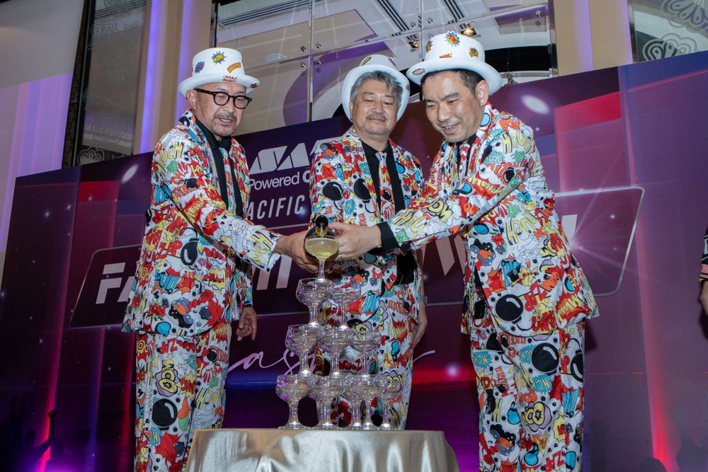Three people wearing matching colorful patterned suits shake hands at an event with purple lighting.