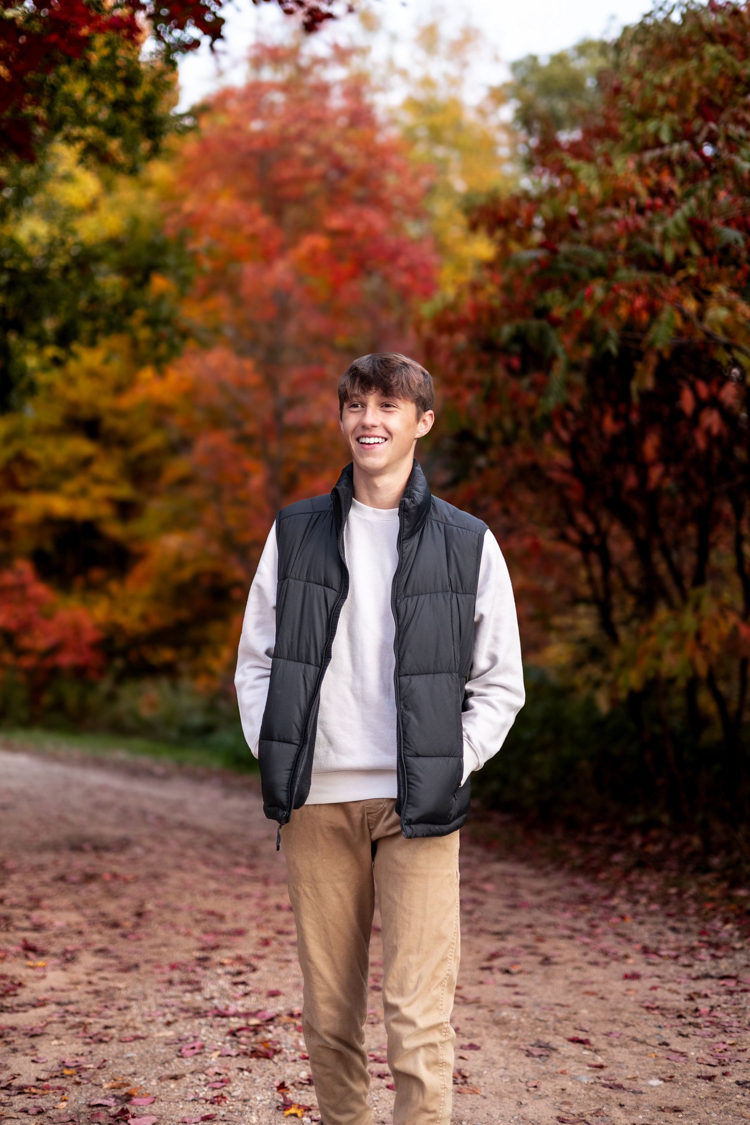 Person wearing a black puffer vest and khaki pants walks on autumn path surrounded by vibrant red and orange fall foliage.