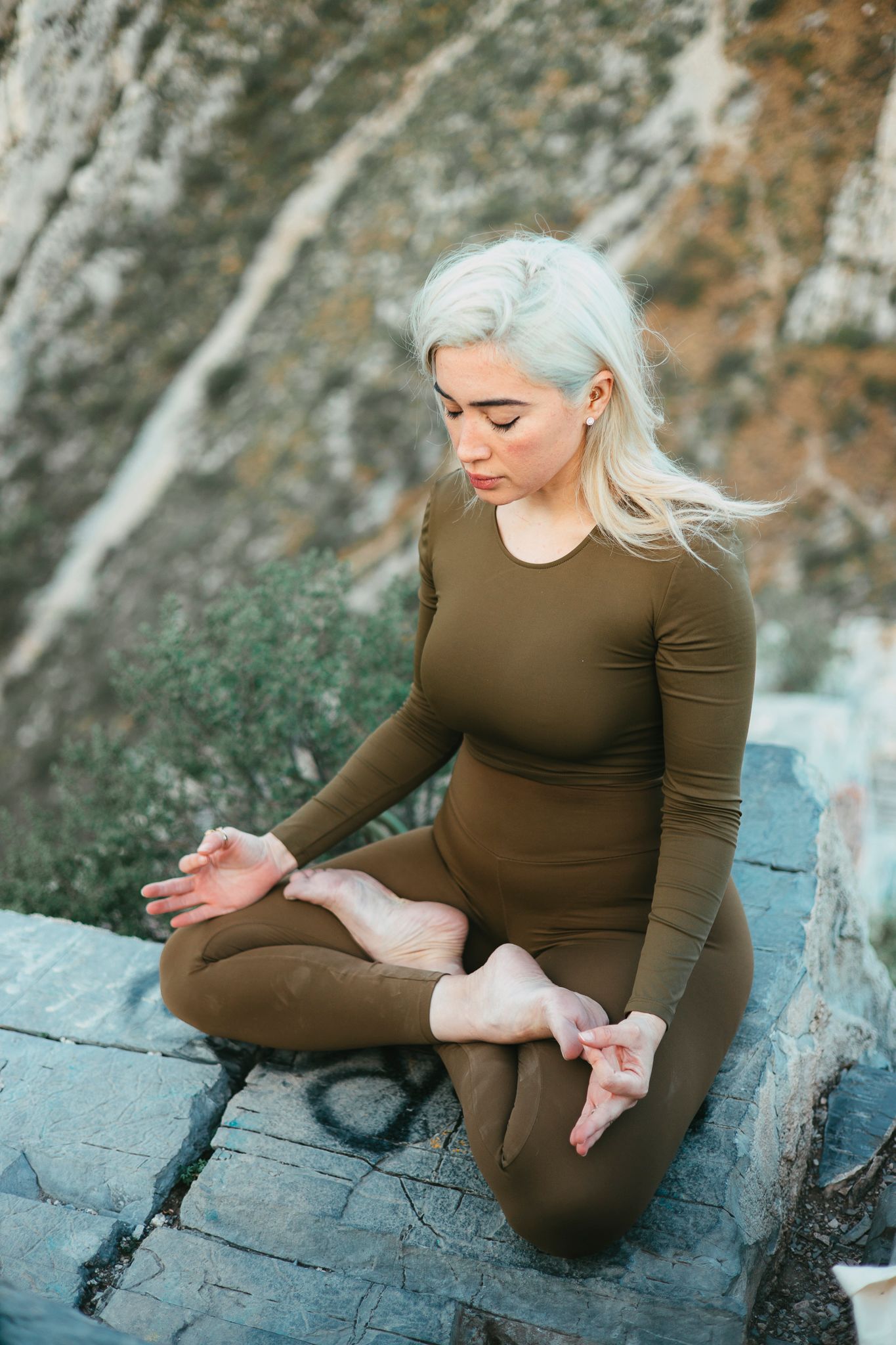 A person meditates in olive-colored athletic wear on rocky terrain with mountains in background.