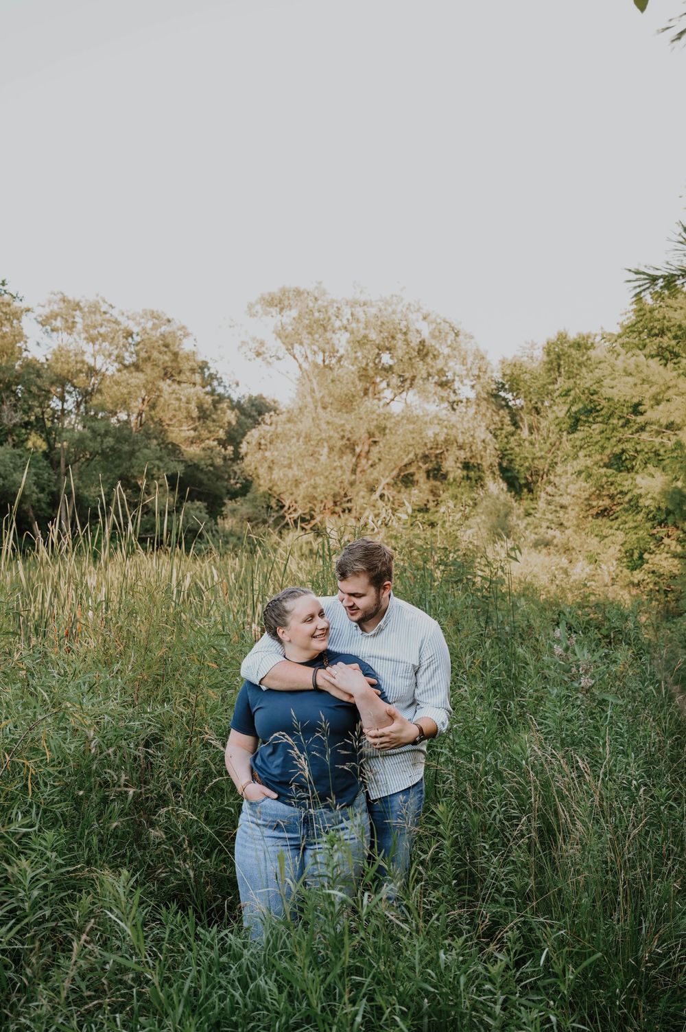 A couple embraces while standing in a tall grass field during golden hour as sunlight filters through trees.