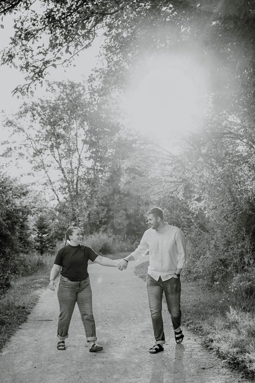 Black and white photo of a couple holding hands while walking together on a tree-lined path outdoors.