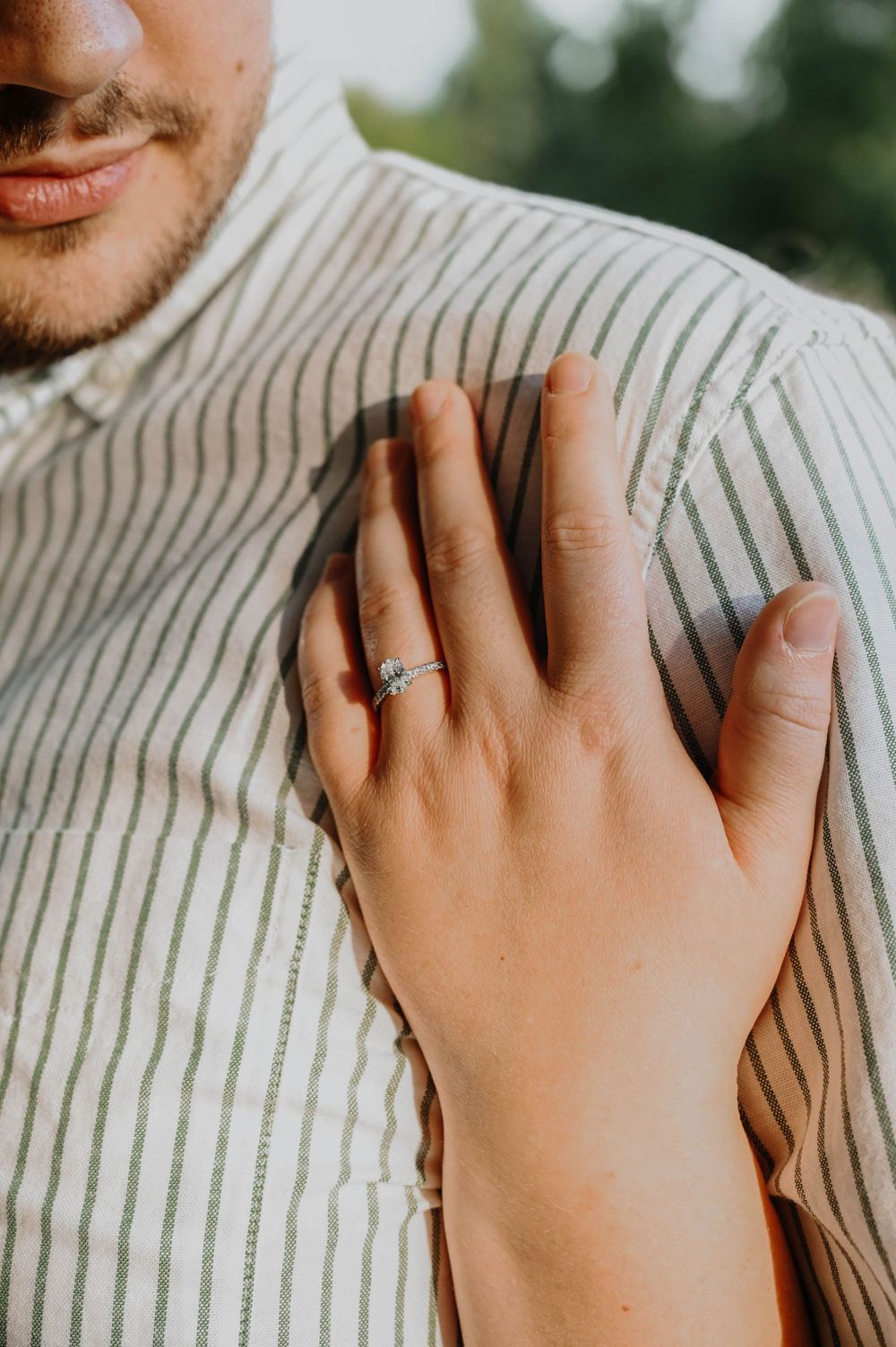 A delicate hand wearing an engagement ring rests on a striped shirt, showing a close-up of the romantic moment.