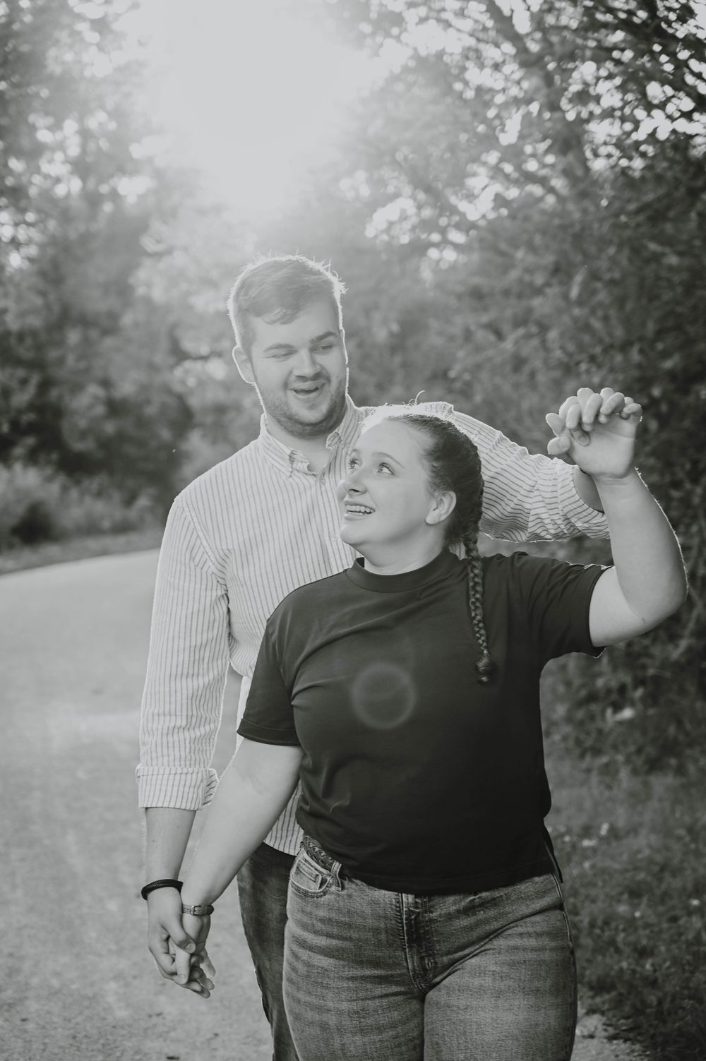 Black and white photo of a loving couple laughing and dancing together on a sunlit path outdoors.