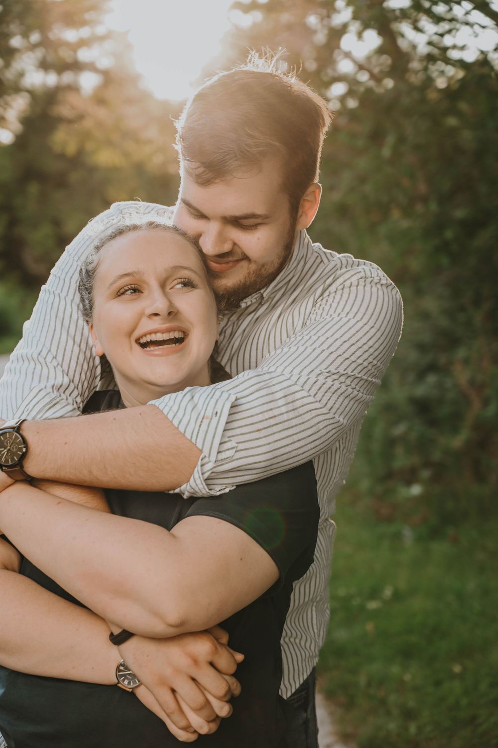 A couple shares a tender embrace and laugh together outdoors during golden hour sunset in a natural setting.