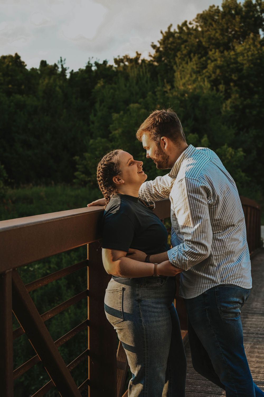 A romantic couple sharing an intimate moment on a wooden bridge at sunset with trees in the background.