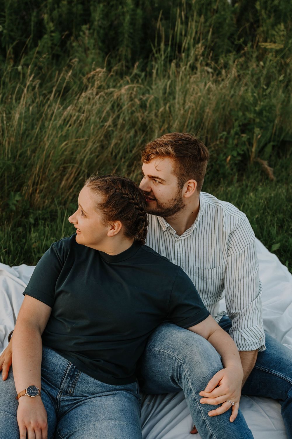 A loving couple sits together on a white blanket outdoors against tall grass and greenery in warm evening light.