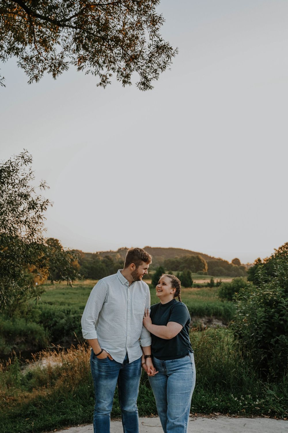 A couple shares a tender moment while standing on a path at sunset with rolling hills and trees in the background.