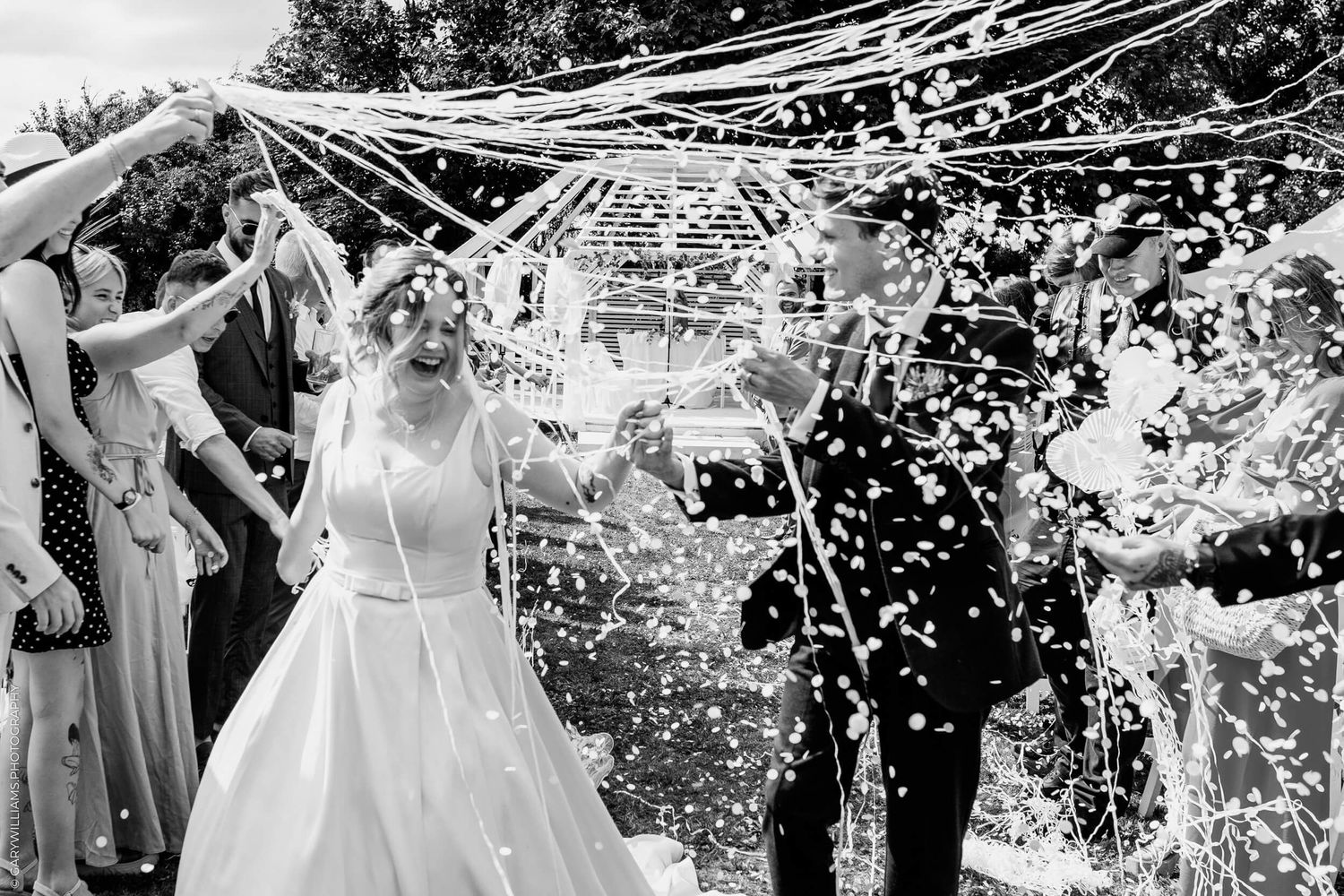 Black and white photo of a wedding celebration with confetti being showered over a white wedding gown under streamers outdoors.