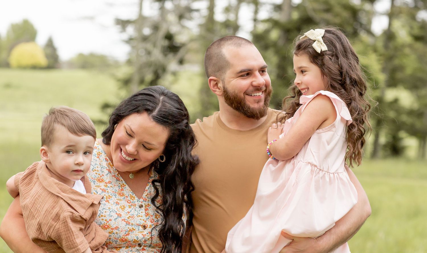Happy family of four smiling outdoors in a lush green park, parents holding young children.