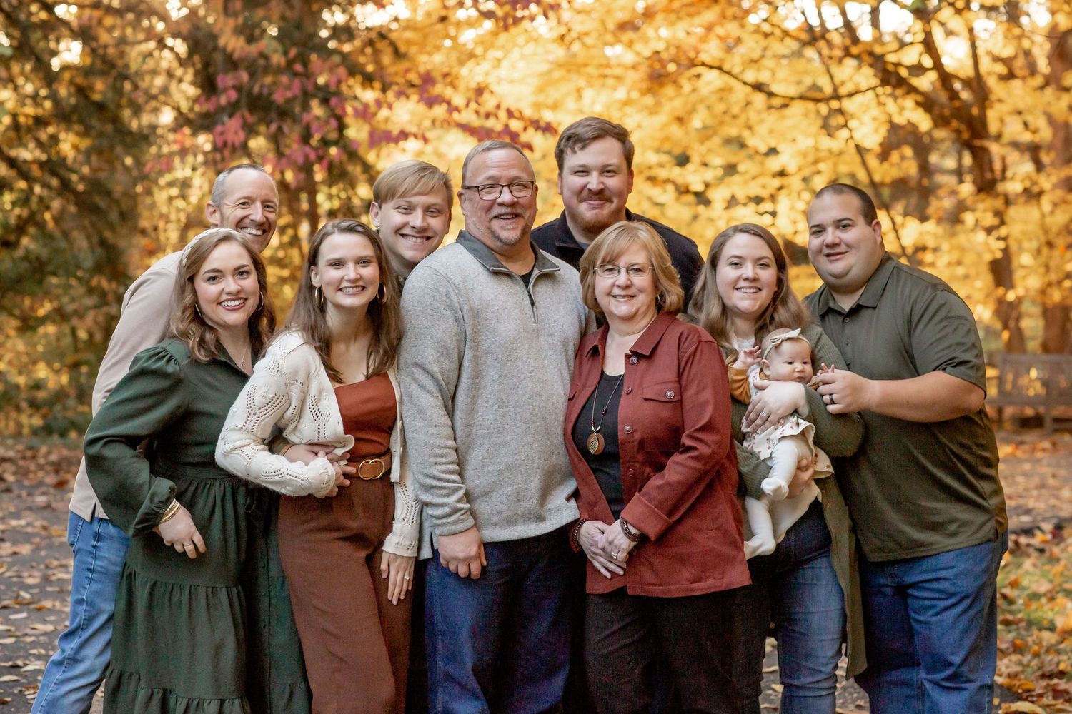 Large family group smiling together surrounded by vibrant fall foliage.
