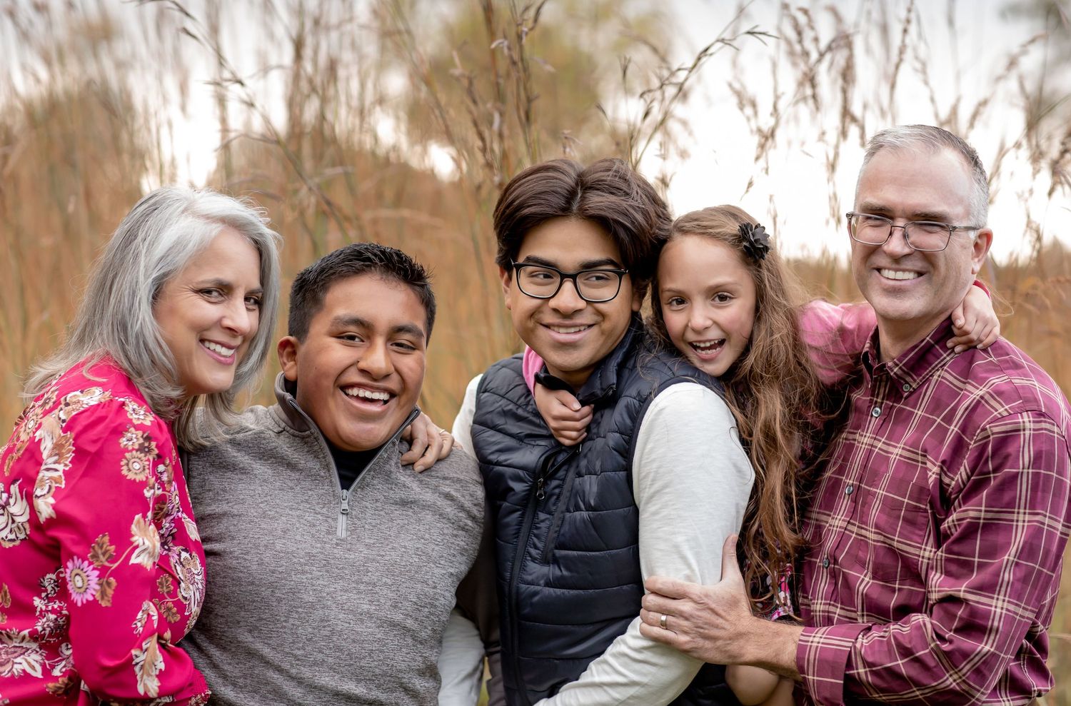 Smiling family of five posing together outdoors with tall grass in background.