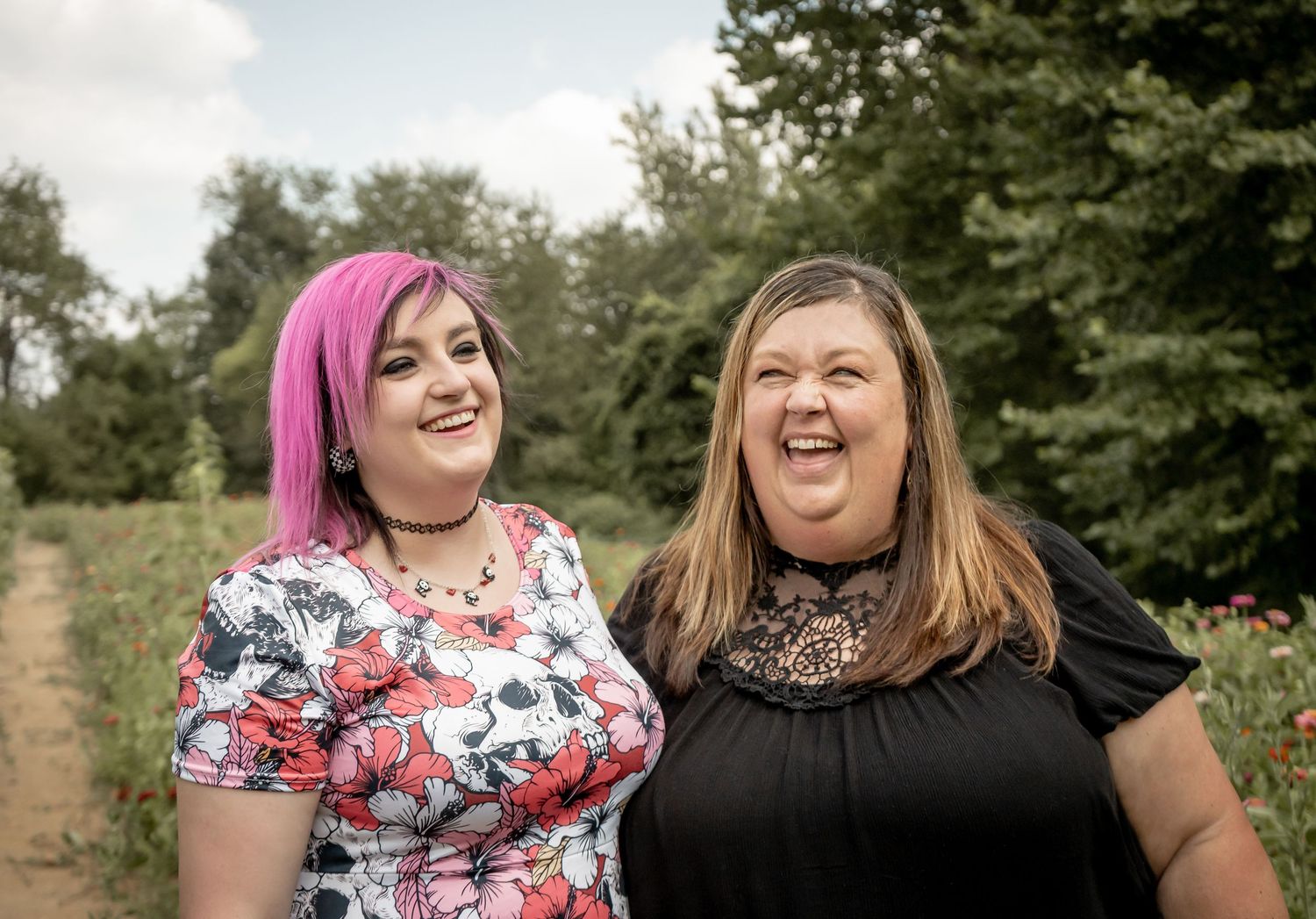 Two women smiling outdoors in a flower field, one with vibrant pink hair in floral dress.