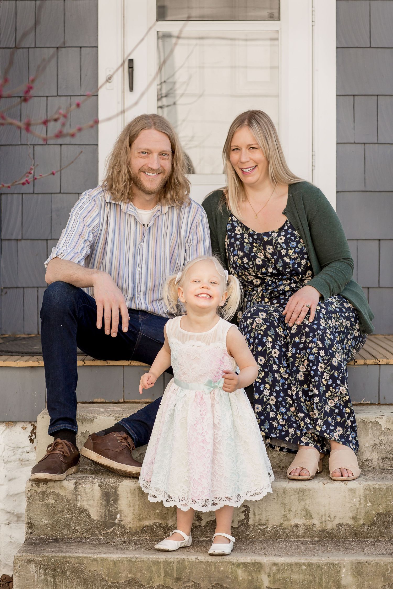 Happy young family with curly-haired father, smiling blonde mother, and toddler daughter in white dress sitting on front porch steps.