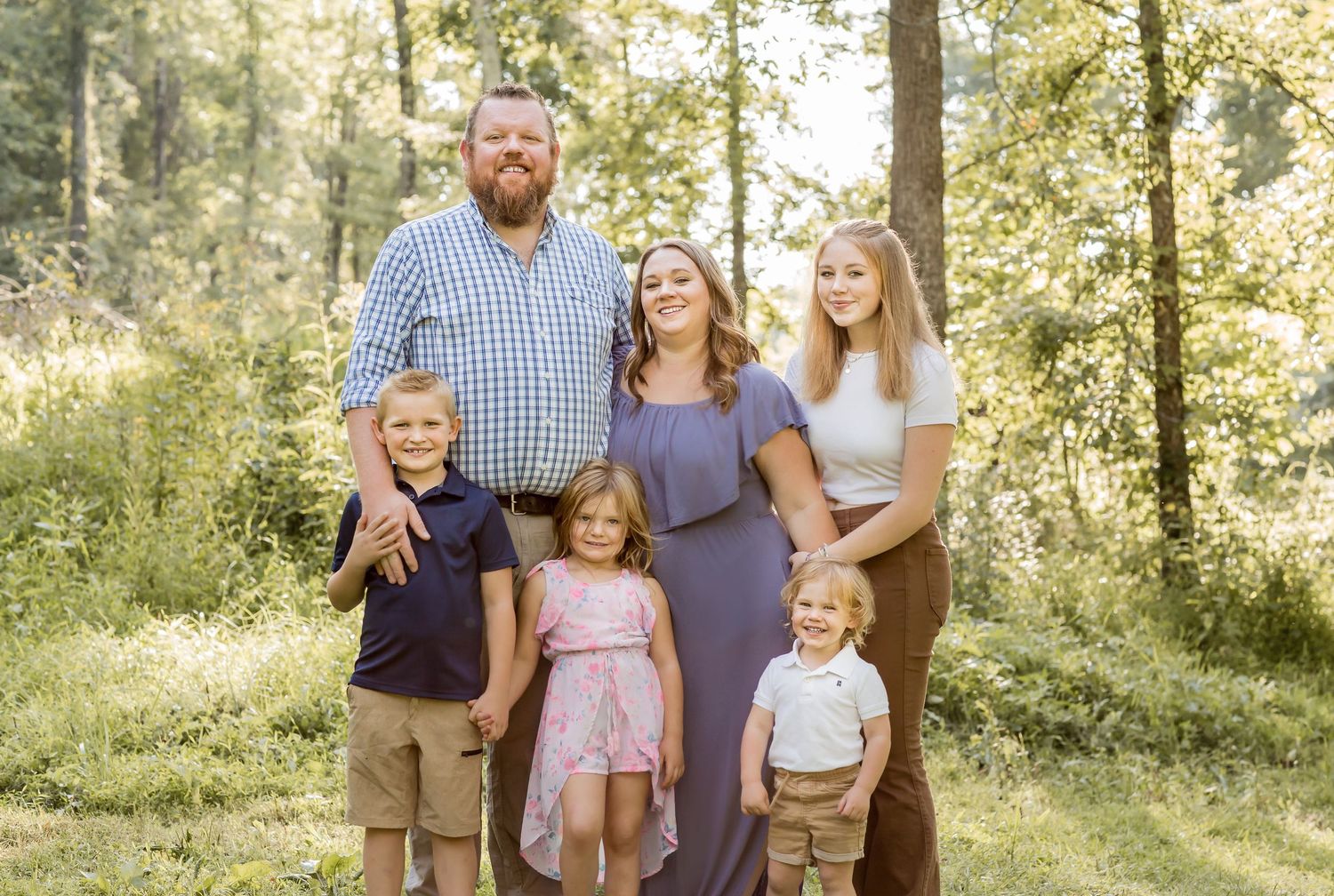 Family of six posing together in a sunny wooded outdoor setting.
