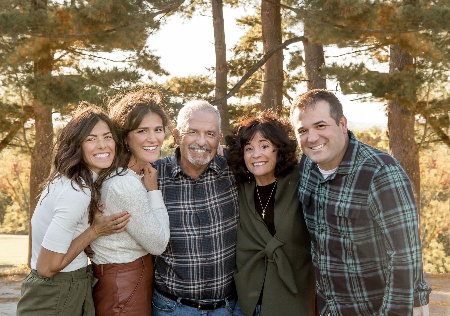 Happy family of five posing together outdoors among tall pine trees in warm sunlight.
