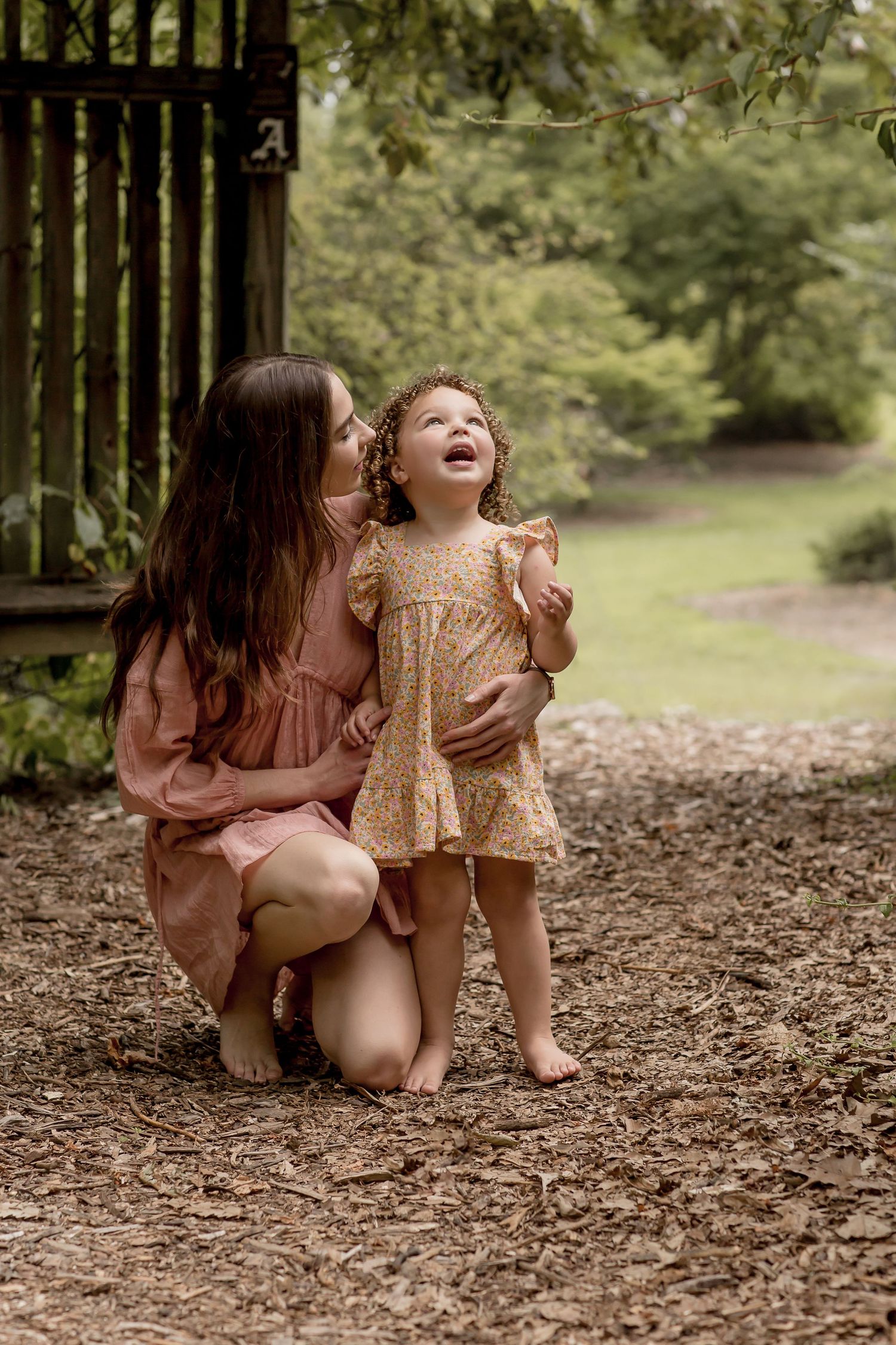 A mother crouches barefoot on wood chips, hugging her laughing toddler daughter in a sunny garden path.