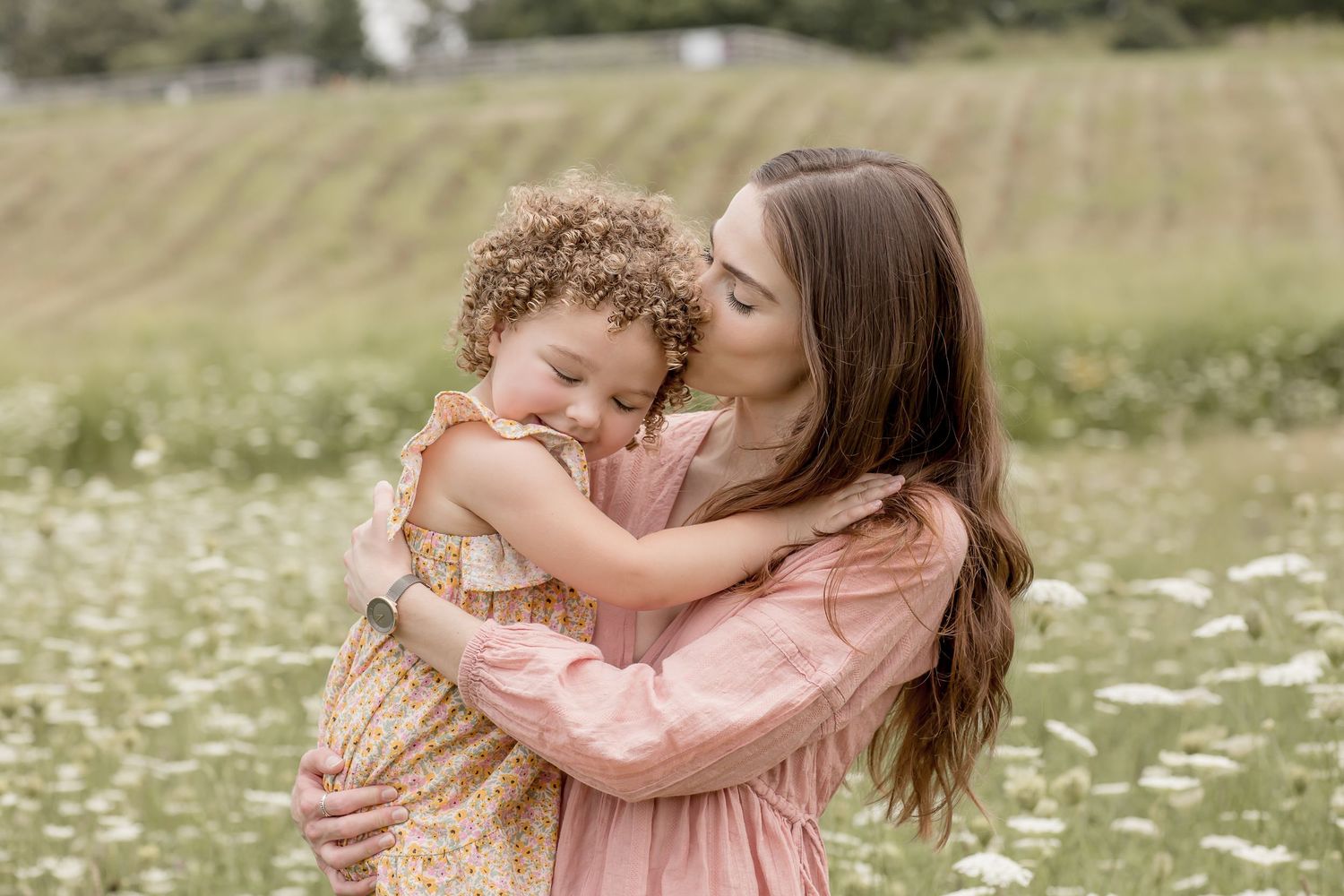 Mother in pink dress kisses curly-haired toddler in flower field on sunny day.