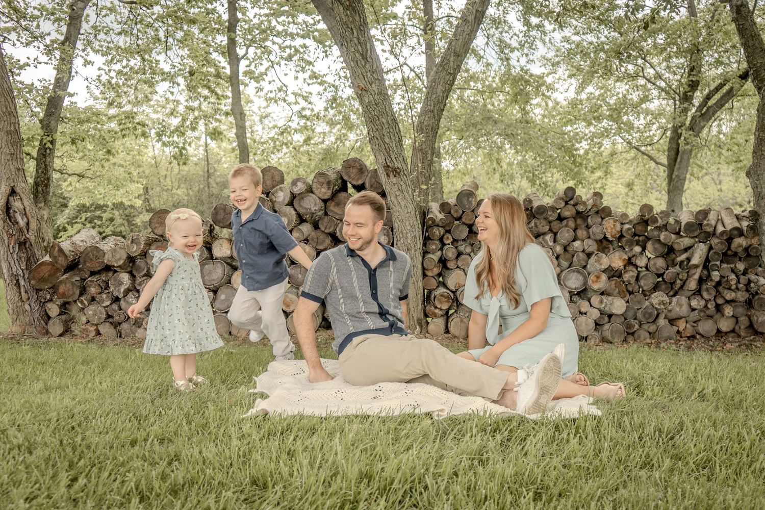 Family of four relaxing on blanket outdoors near stacked logs, two young children playing around parents.