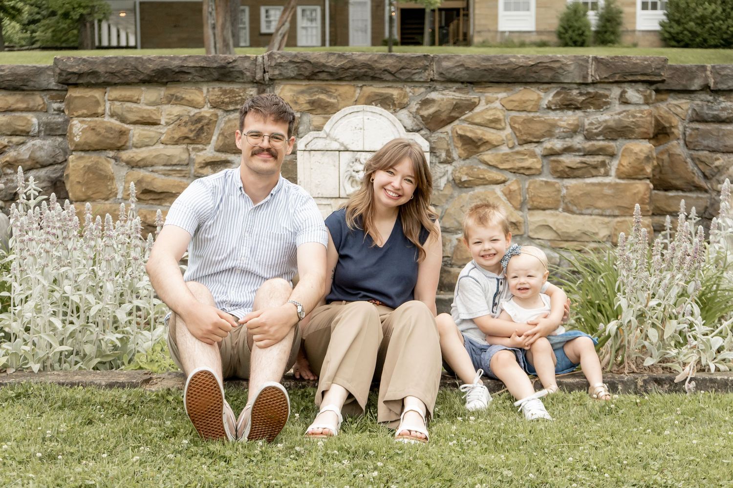 Family of four sitting on green lawn in front of stone wall on sunny day.