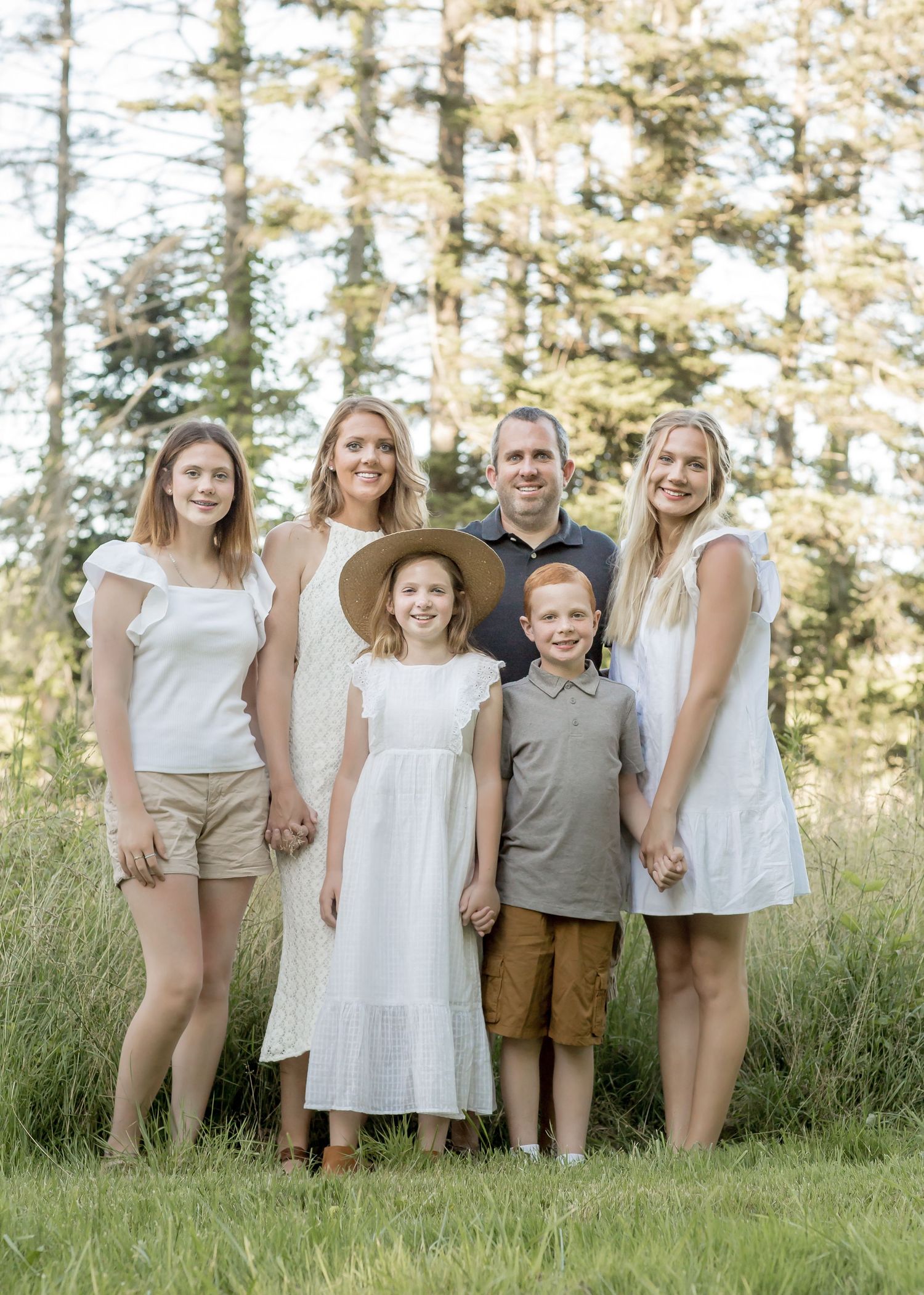 A family of six in coordinating white and neutral outfits poses in a grassy meadow surrounded by tall trees.