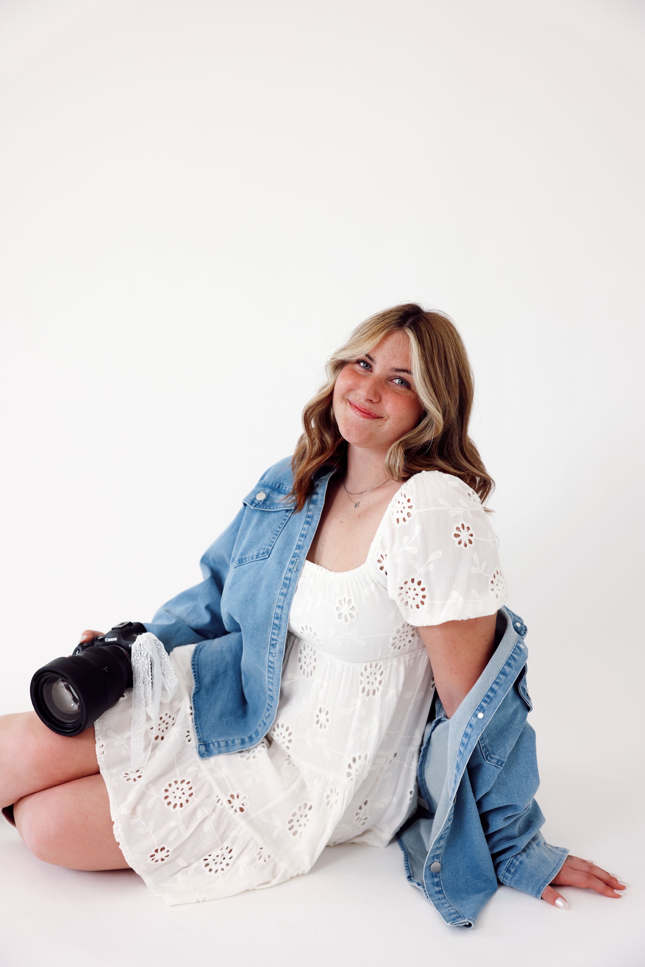 Professional photographer in denim overalls and white sweater sits casually on white blocks holding camera against studio backdrop.