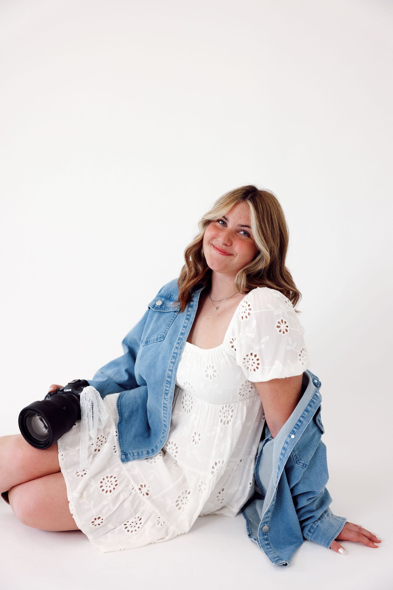 Professional photographer in denim overalls and white sweater sits casually on white blocks holding camera against studio backdrop.