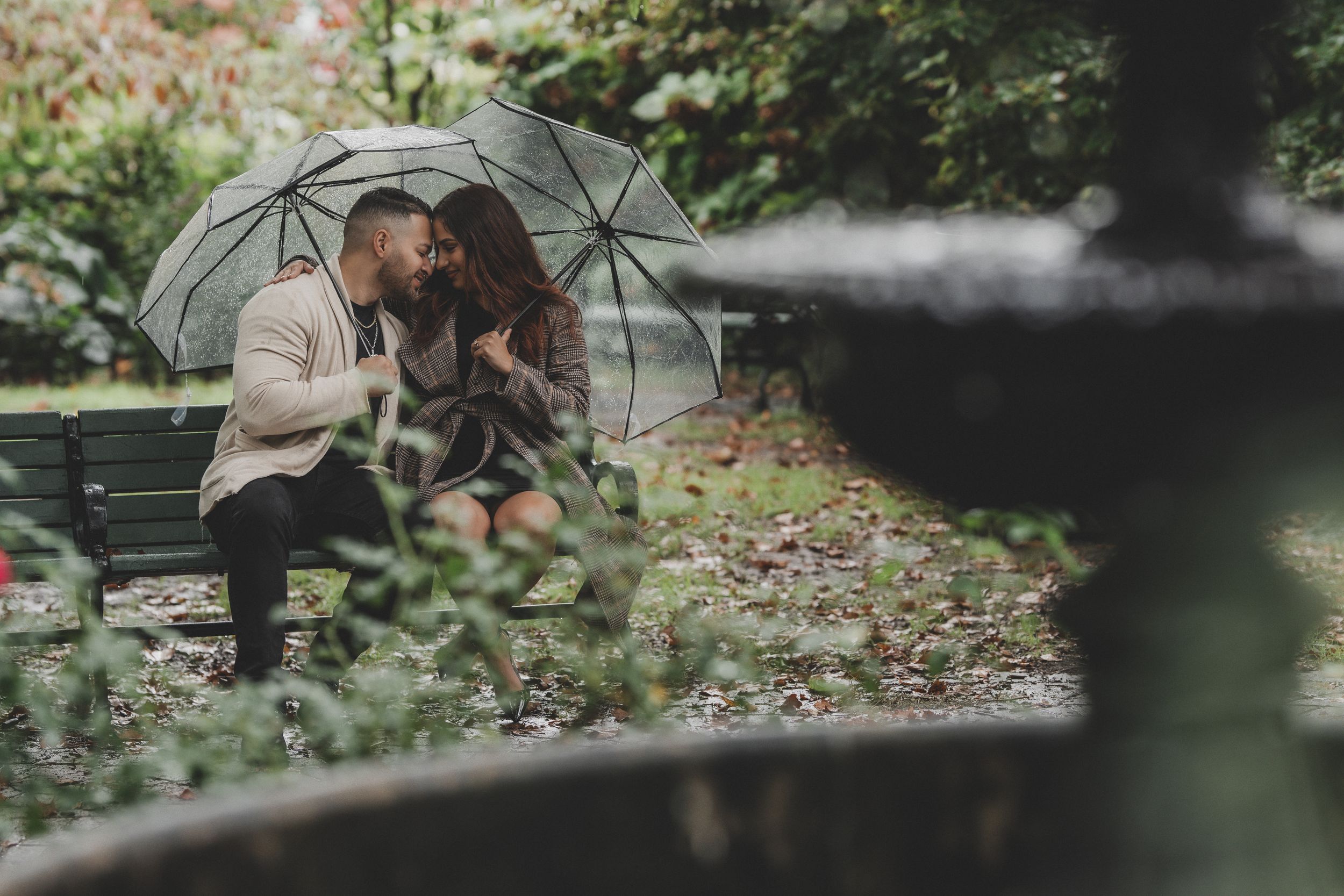 A couple shares a romantic moment under a clear umbrella on a park bench surrounded by autumn leaves.