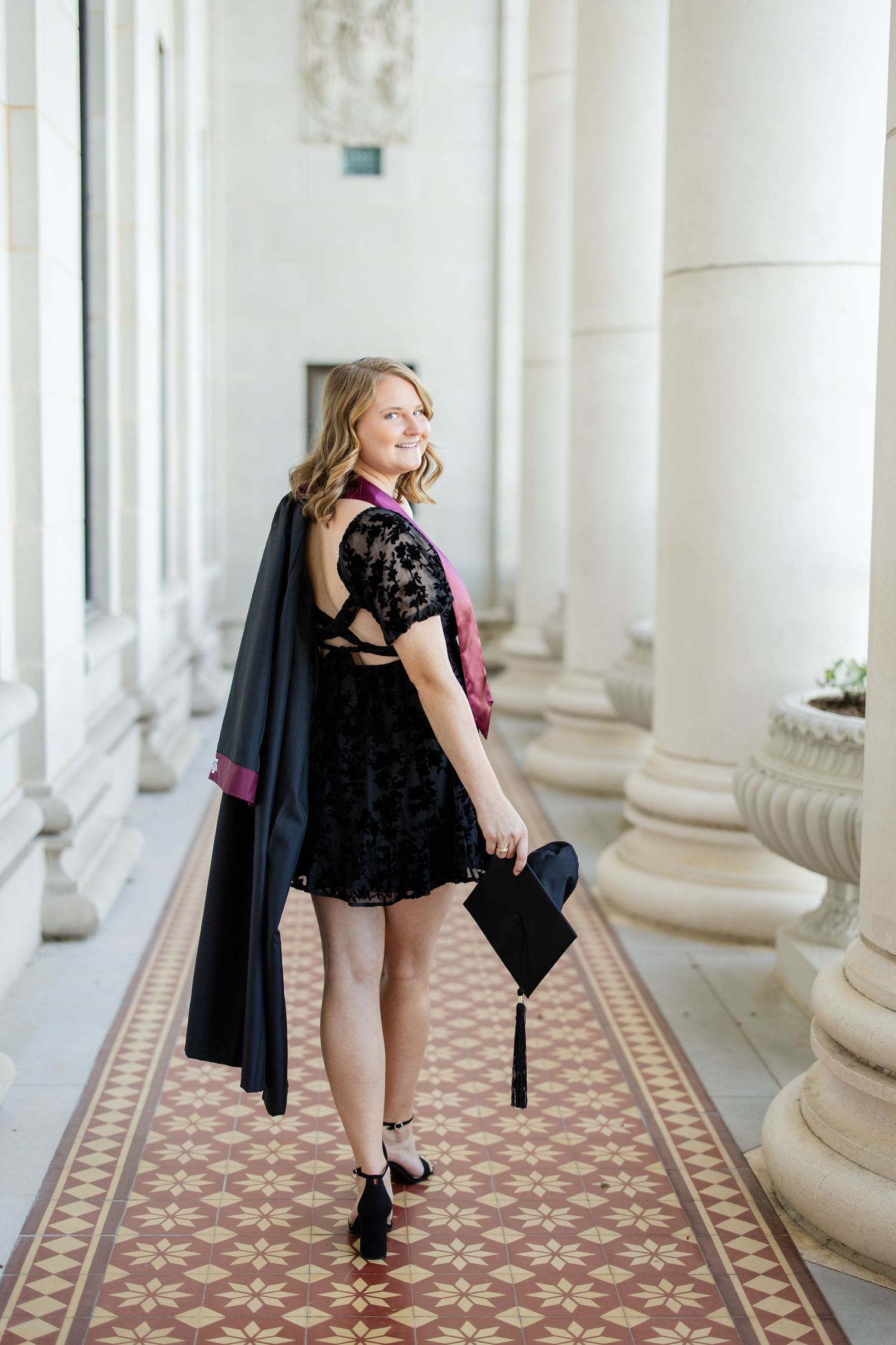 Graduate in black lace dress and graduation gown poses in ornate hallway with decorative columns and patterned floor.