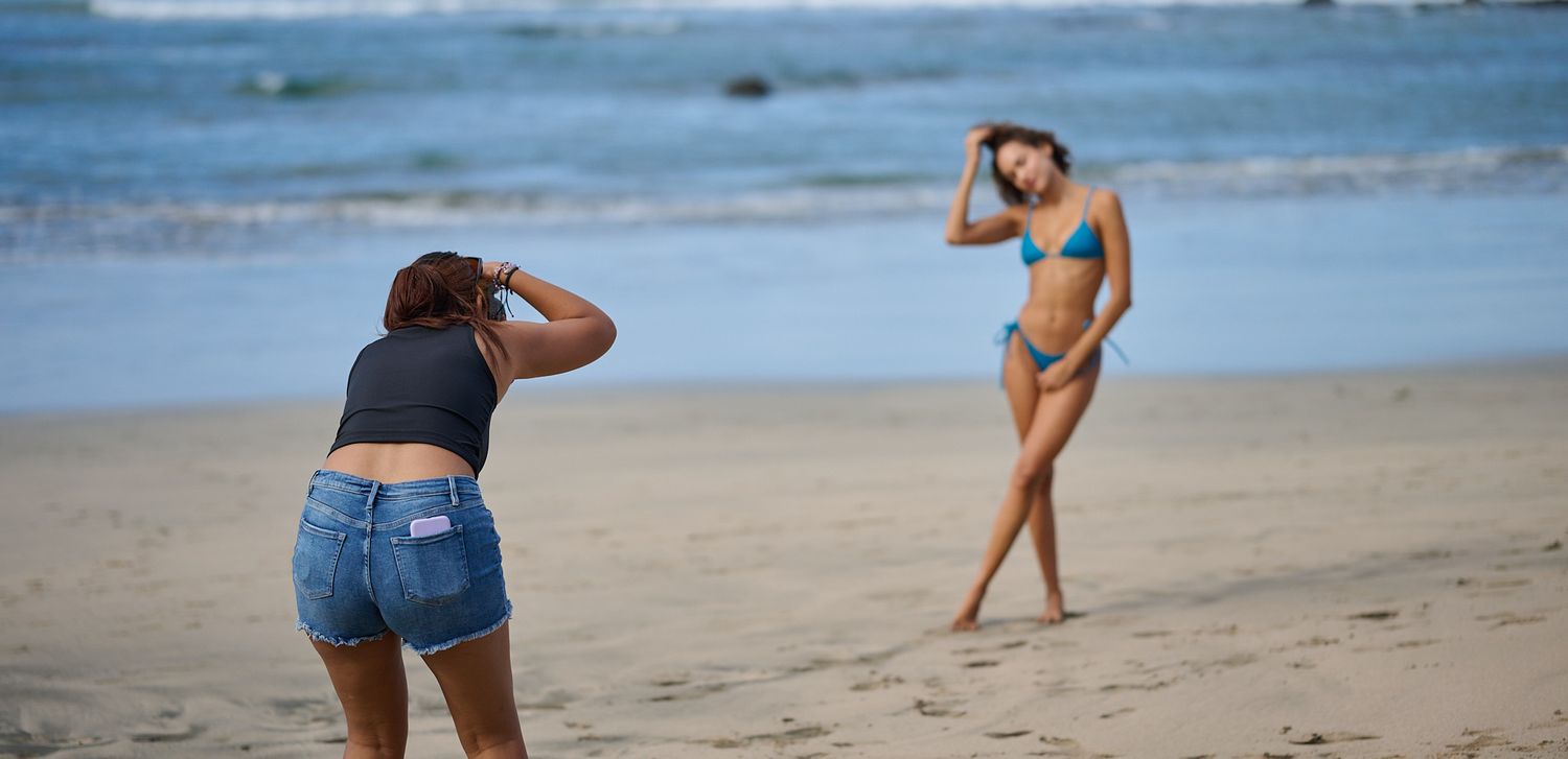 A photographer takes photos of a model posing on a sandy beach with blue ocean waves in the background.
