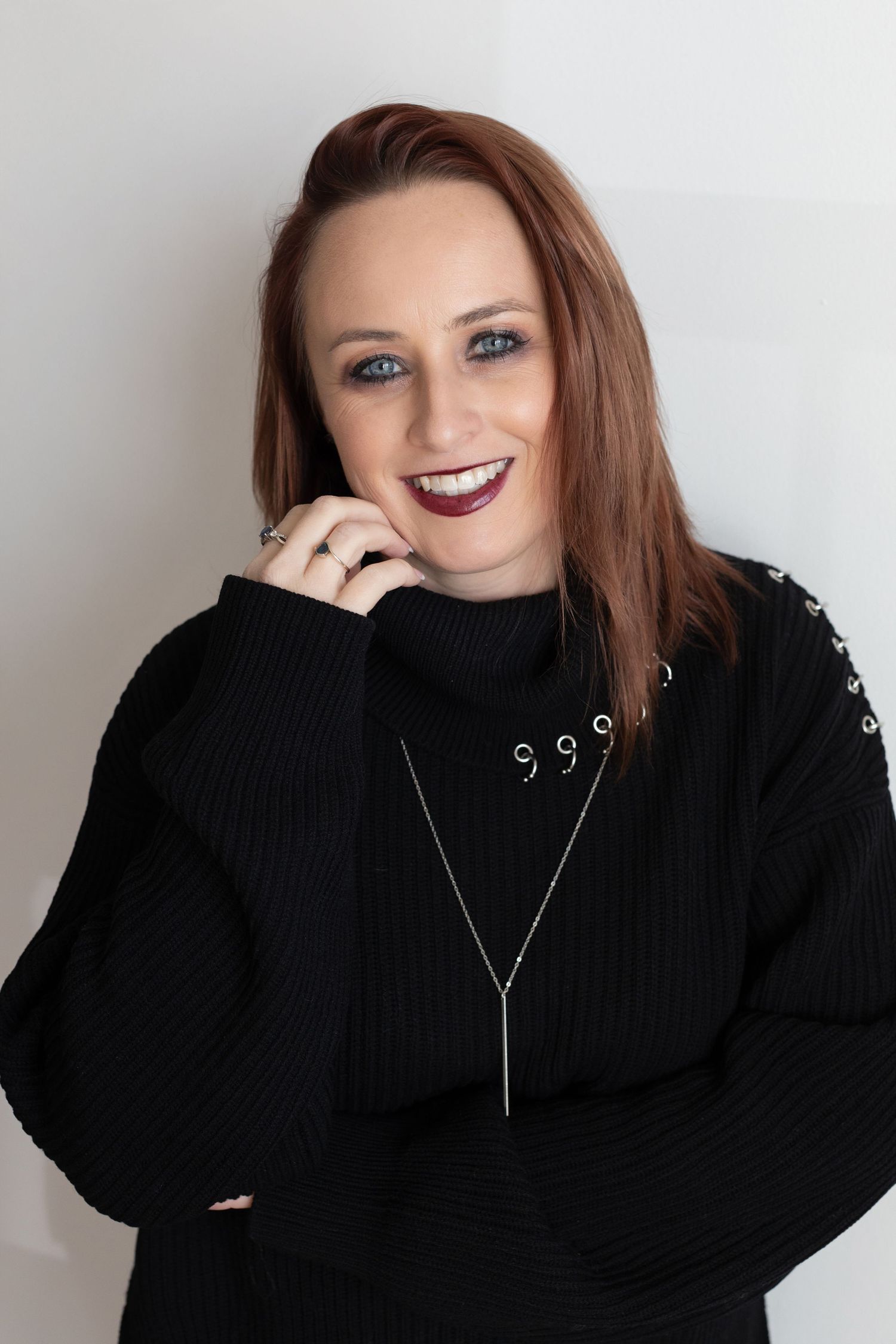 Portrait of someone with auburn hair wearing a black ribbed sweater and silver necklace against a light background.